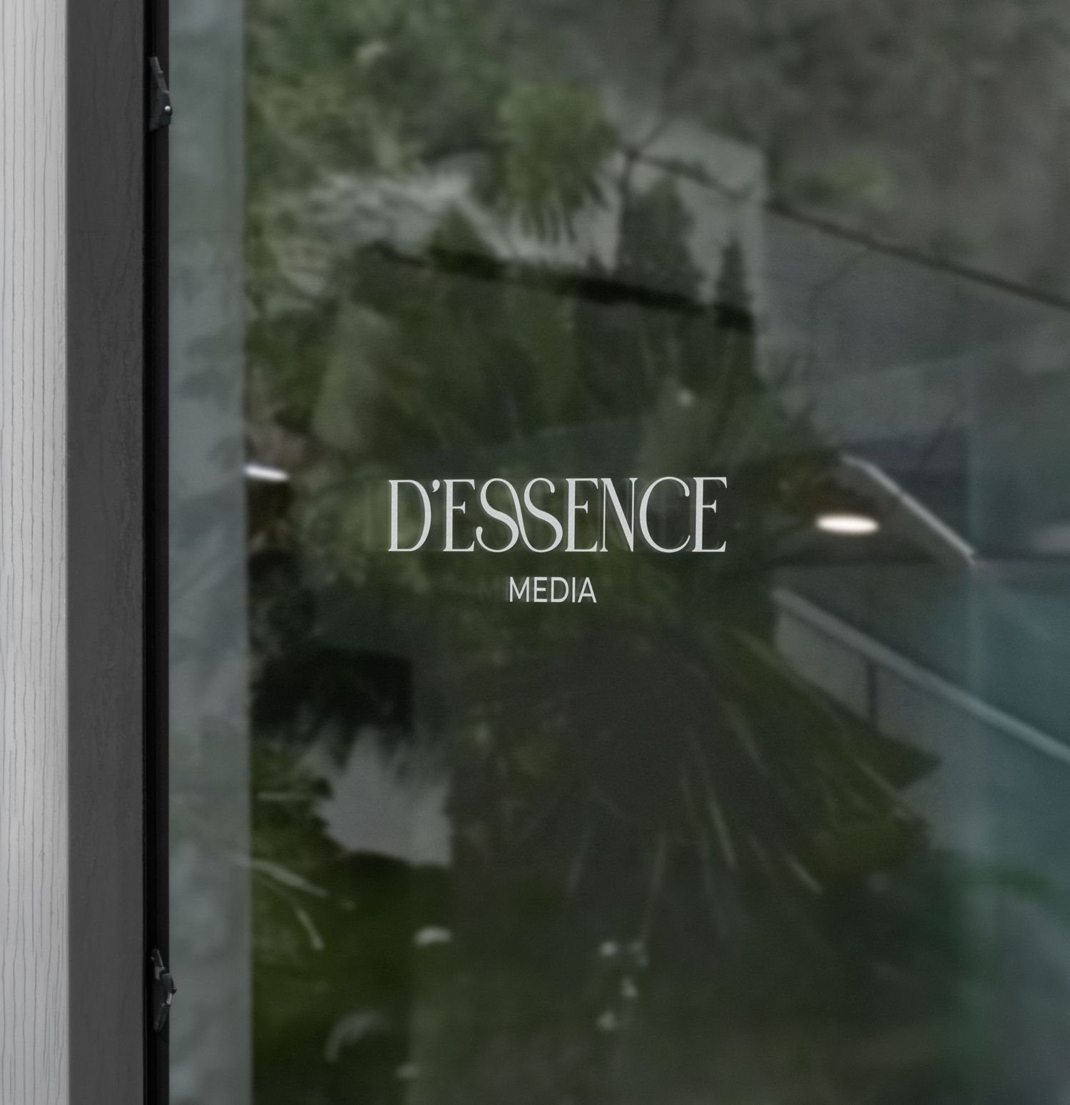 Dessence Media logo reflected on glass window with green plants visible in background.