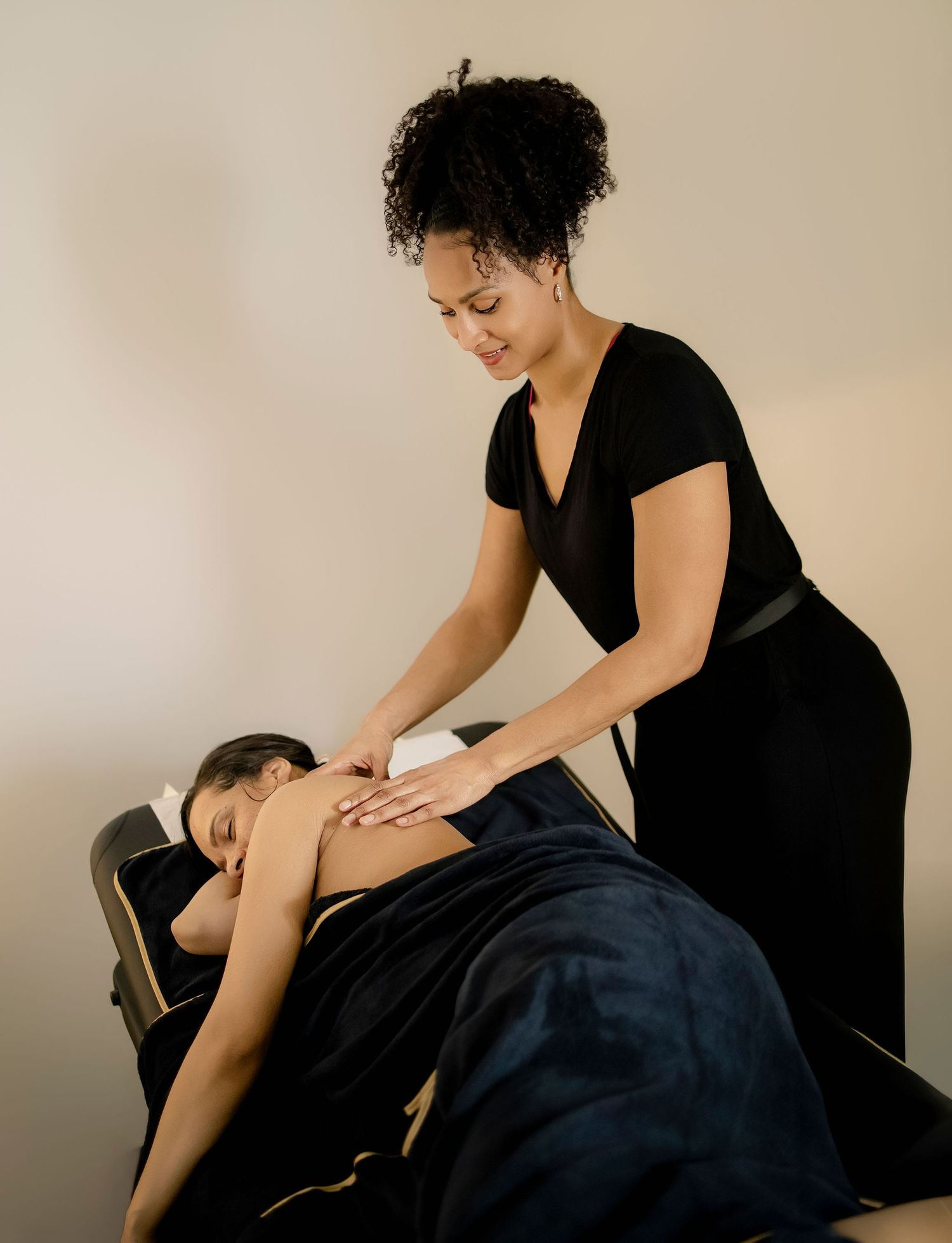 A massage therapist in black attire performs a relaxing back massage on a client lying on a massage table.
