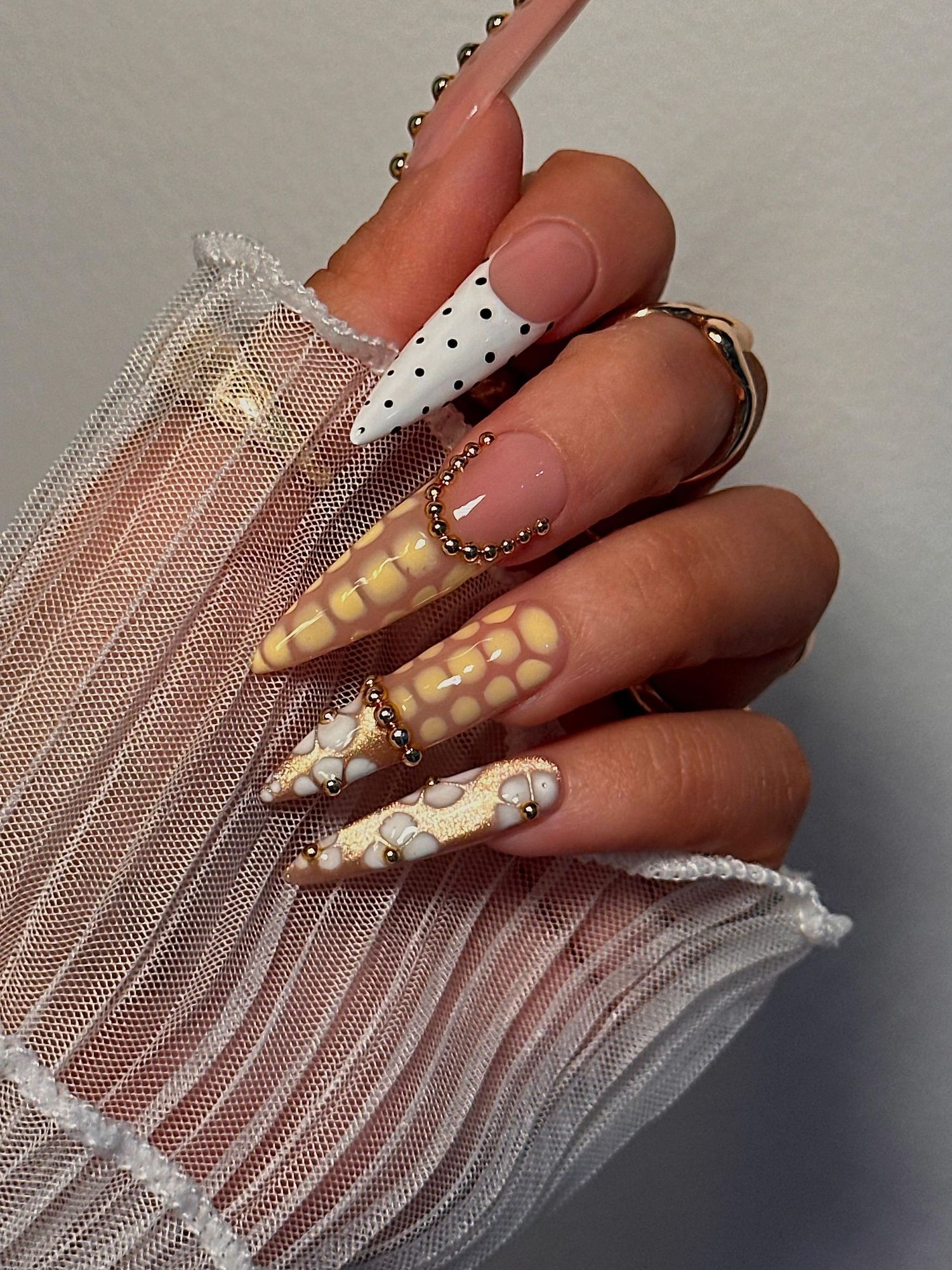Elegant stiletto nails featuring gold snake print, polka dots, and French tips adorned with gold beaded rings.