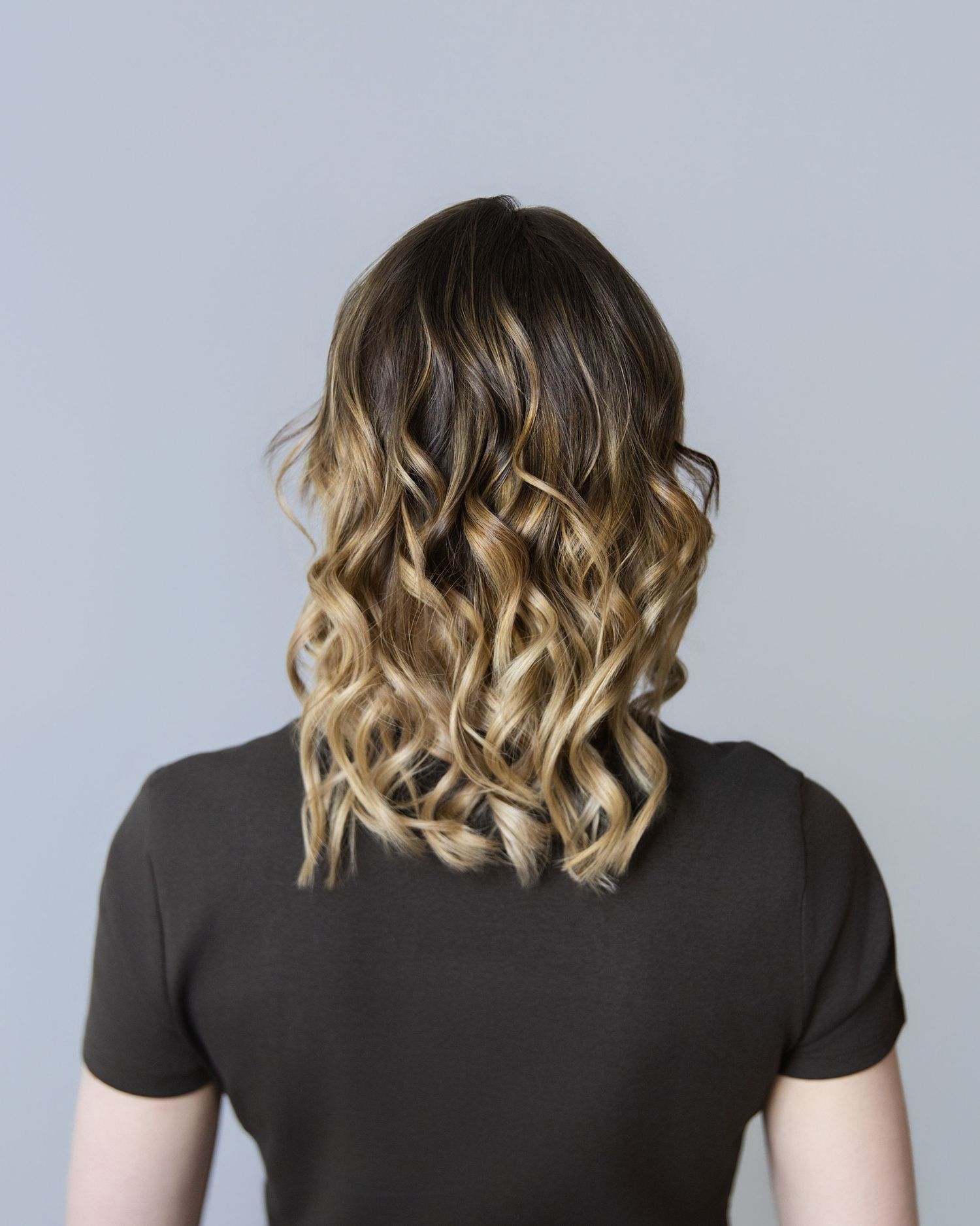 Back view of a woman with ombre balayage hair, transitioning from dark brown roots to blonde curly ends.