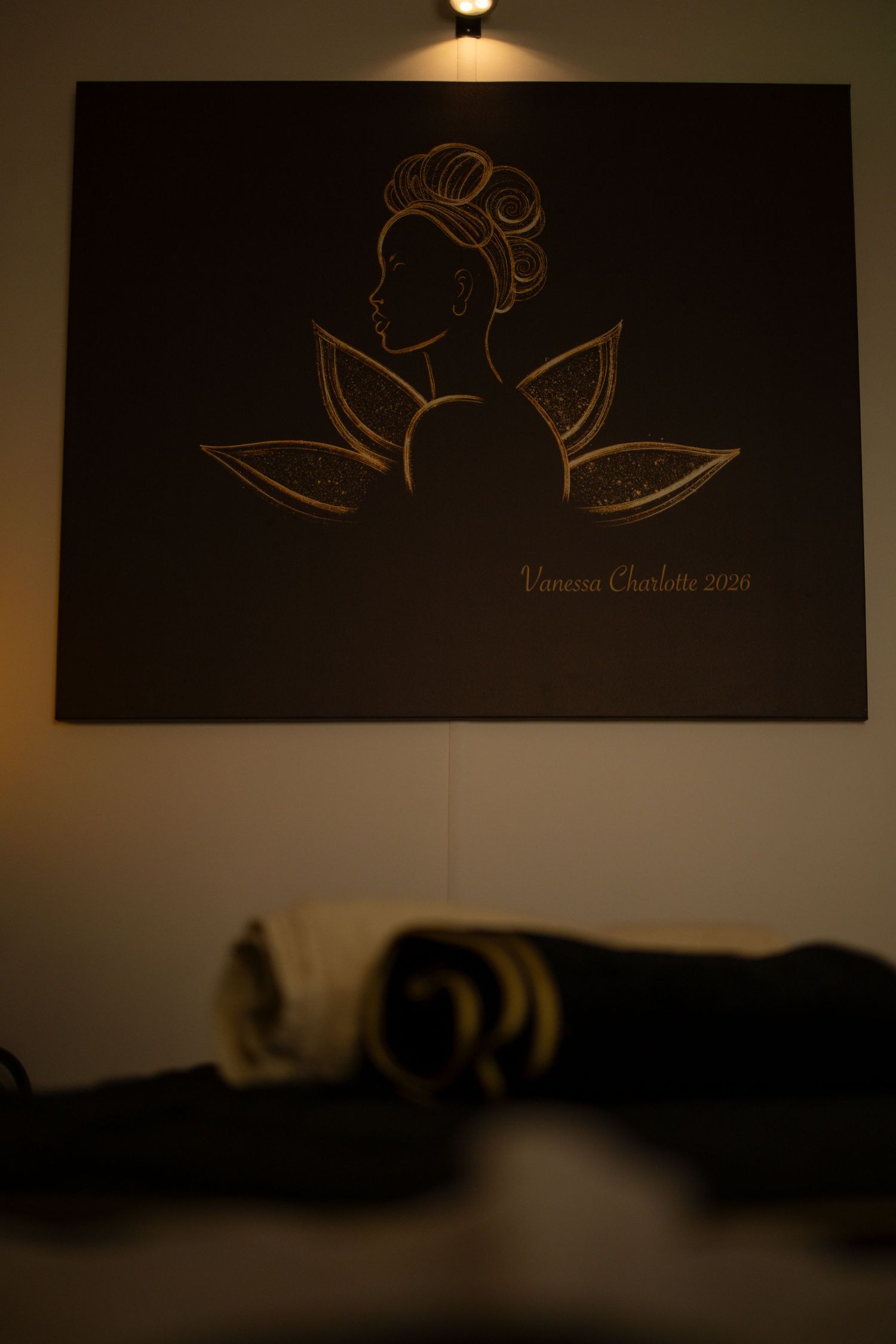 Dark spa room with golden logo artwork by Vanessa Charlotte 2020 featuring woman silhouette, with rolled towels in foreground.