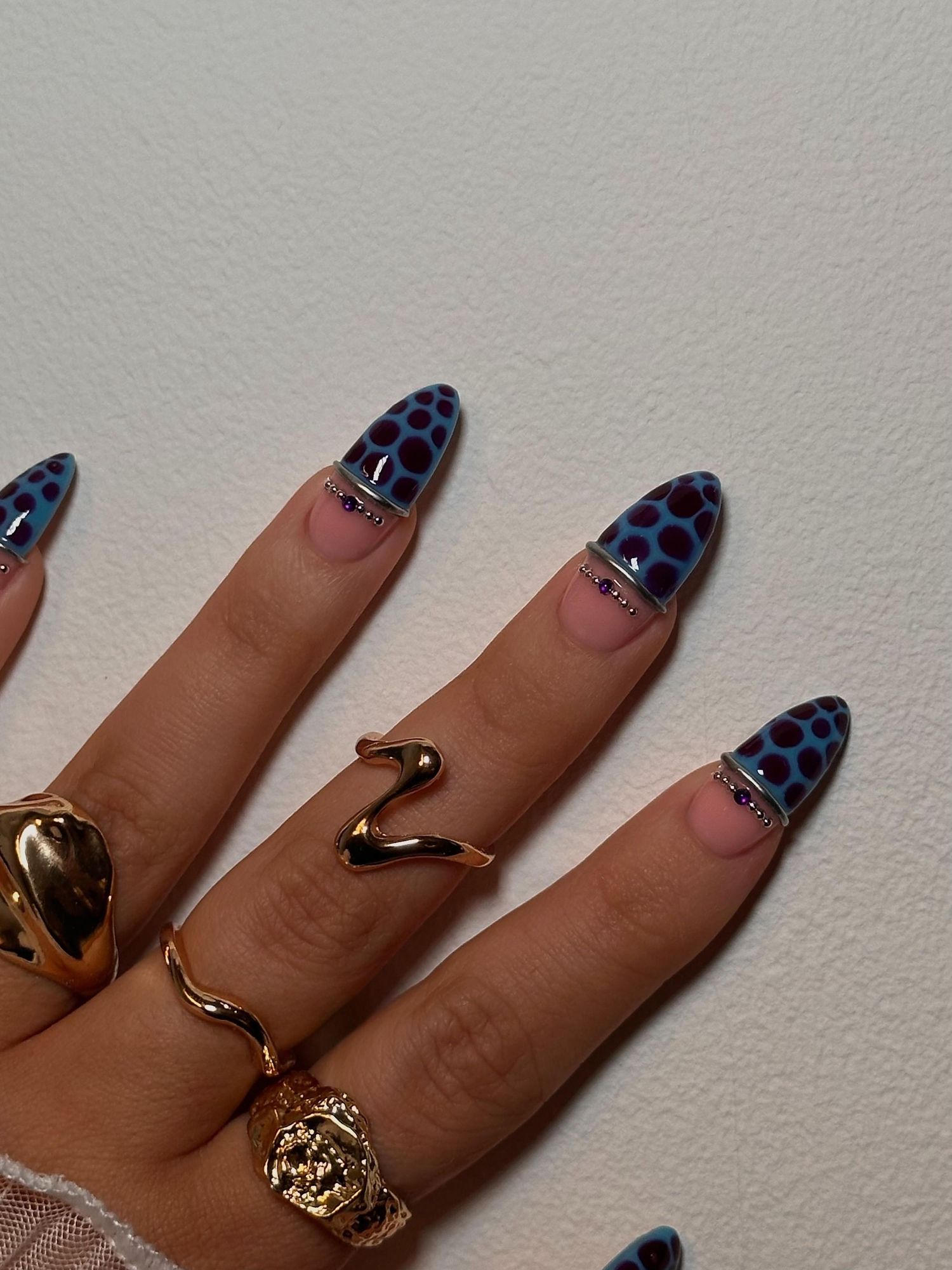 Hand with almond-shaped nails featuring blue leopard print nail art, adorned with gold snake and lion rings.