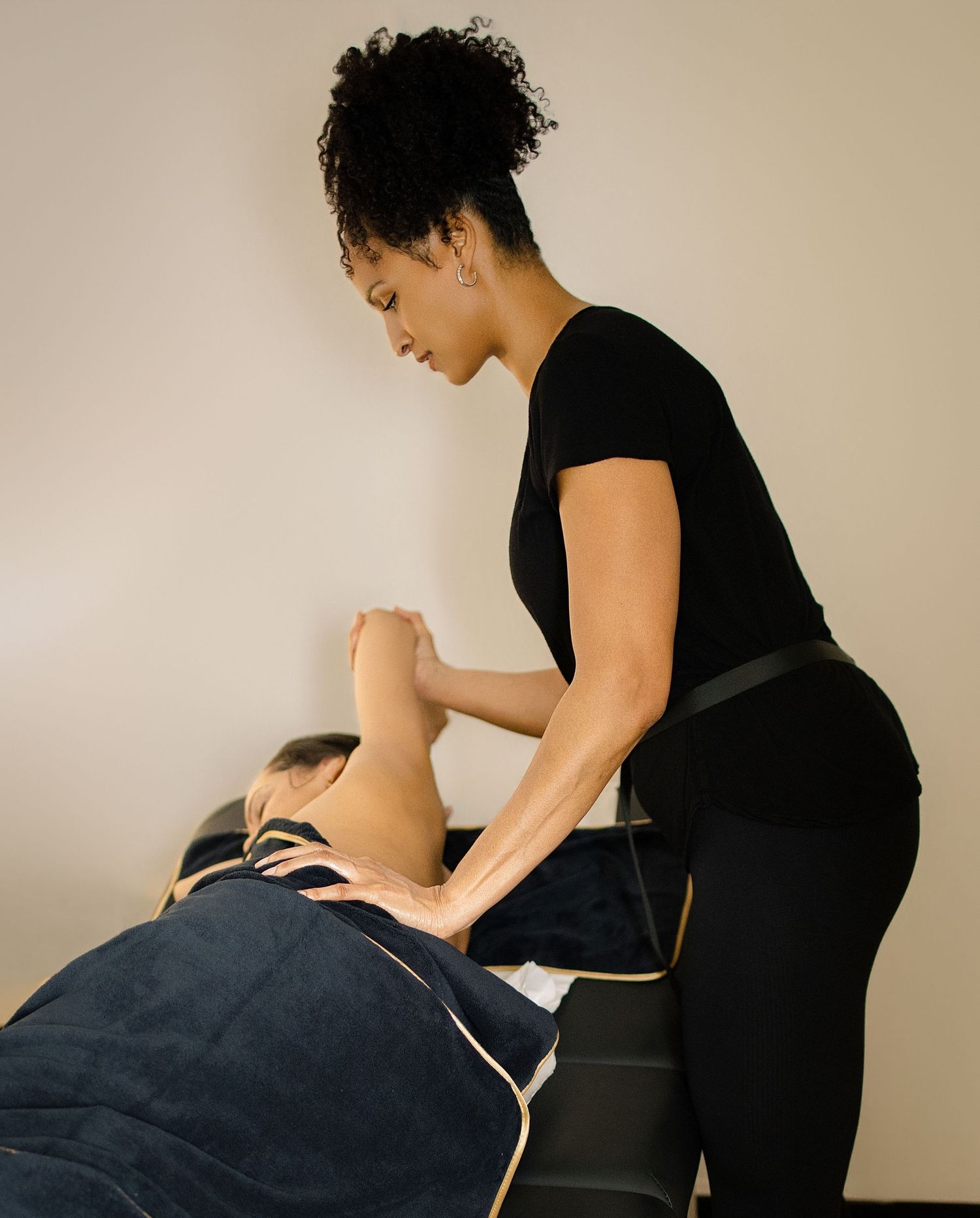 A massage therapist in black attire performs a professional arm massage on a client lying on a treatment table.