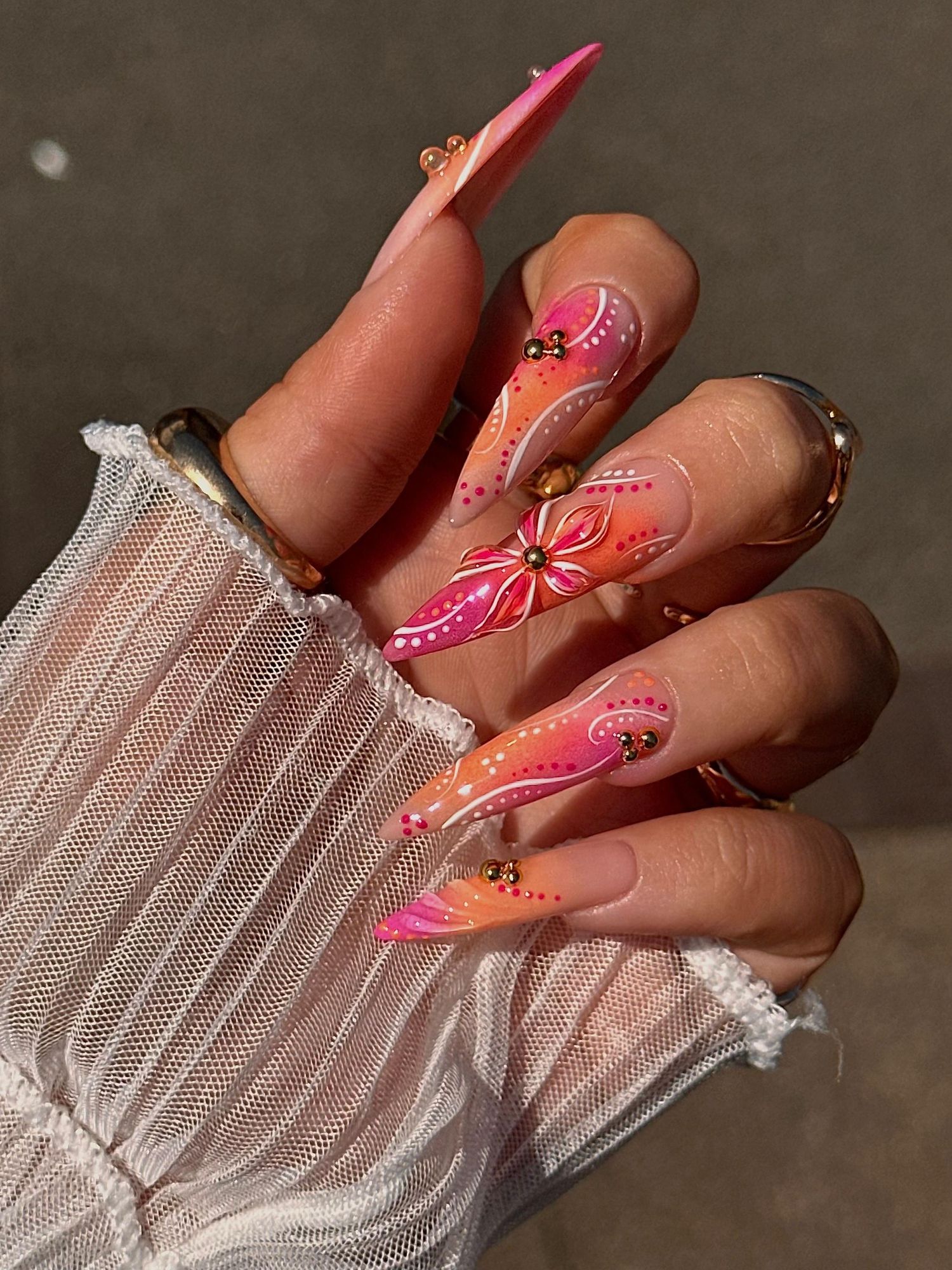Long stiletto nails with pink-orange ombre, floral designs, swirls, and rhinestone accents on manicured hands.