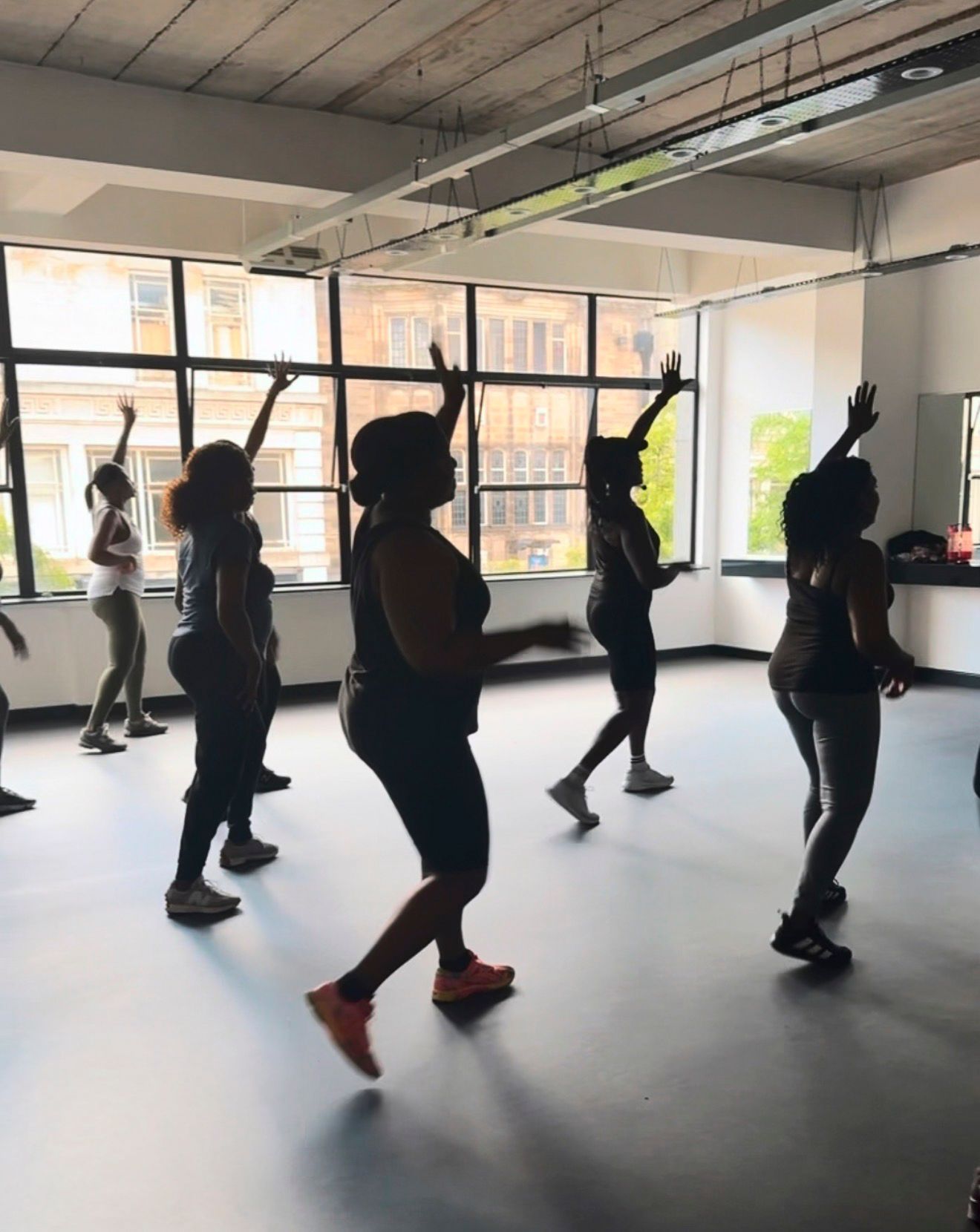 Group of dancers mid-movement in a bright studio, arms raised, silhouetted against large windows.