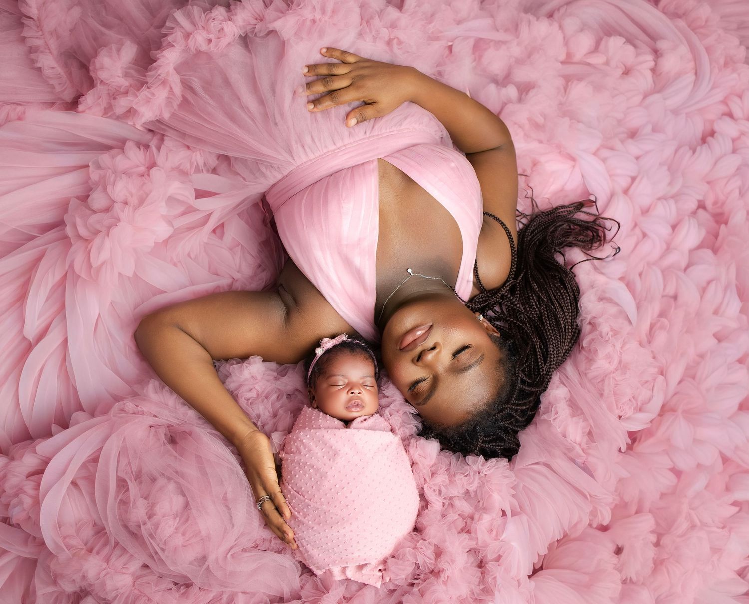 A Black mother in a pink dress lies with her swaddled newborn on a fluffy pink tulle backdrop.