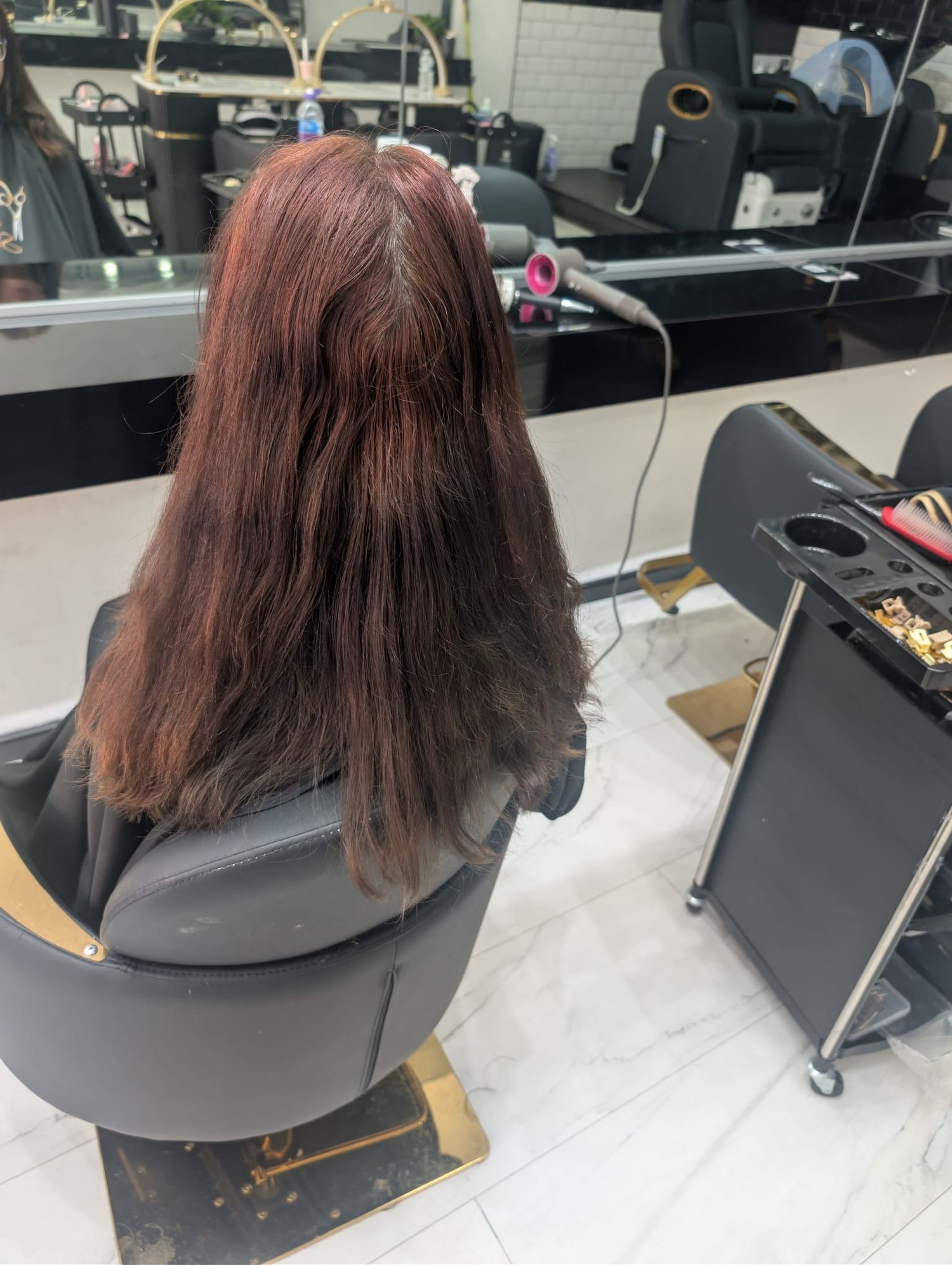 Woman with long reddish-brown hair sitting in salon chair, viewed from behind in a professional hair salon.