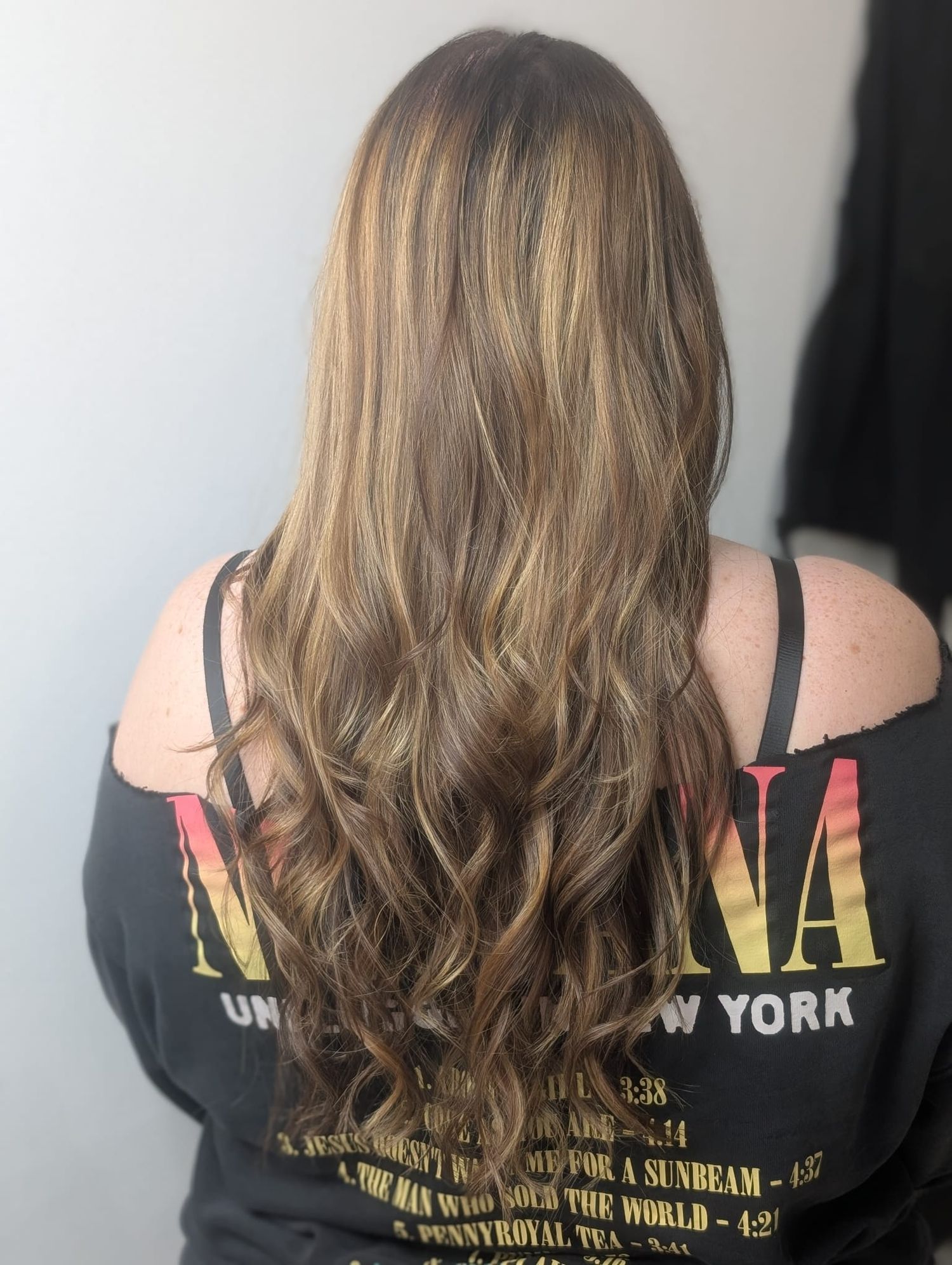Woman with long wavy brown hair featuring blonde highlights, wearing a black graphic tee, viewed from behind.