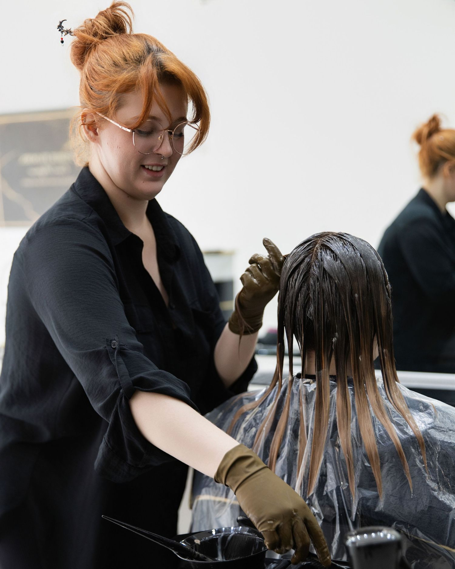 A smiling redhead hairstylist applies color treatment to a client's hair while wearing black gloves in a salon.