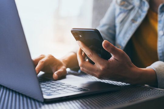 Person using a smartphone while working on a laptop at a desk in a bright, modern workspace.
