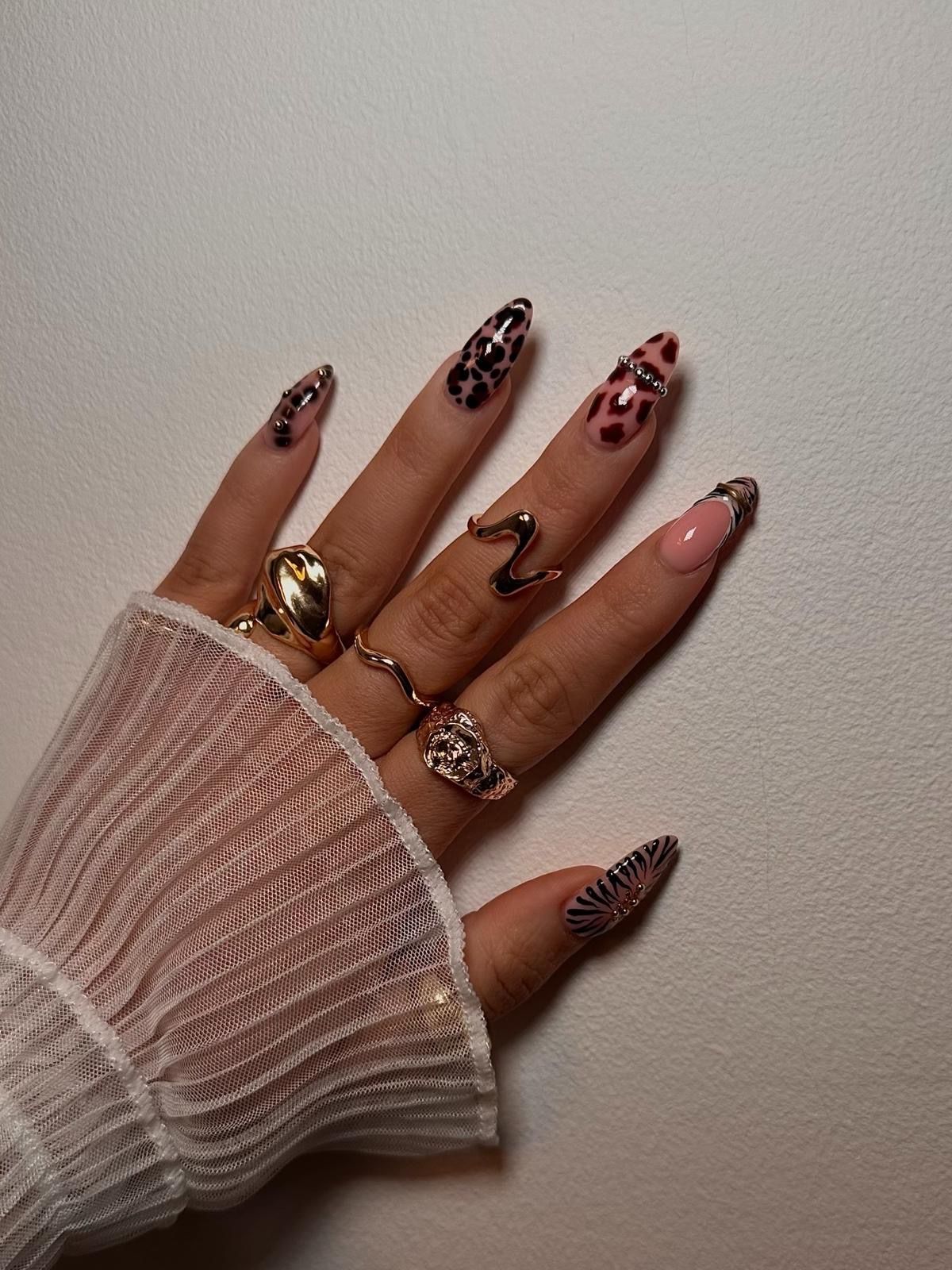 Hand with long almond nails featuring leopard print, wavy lines, and floral designs, adorned with gold rings.