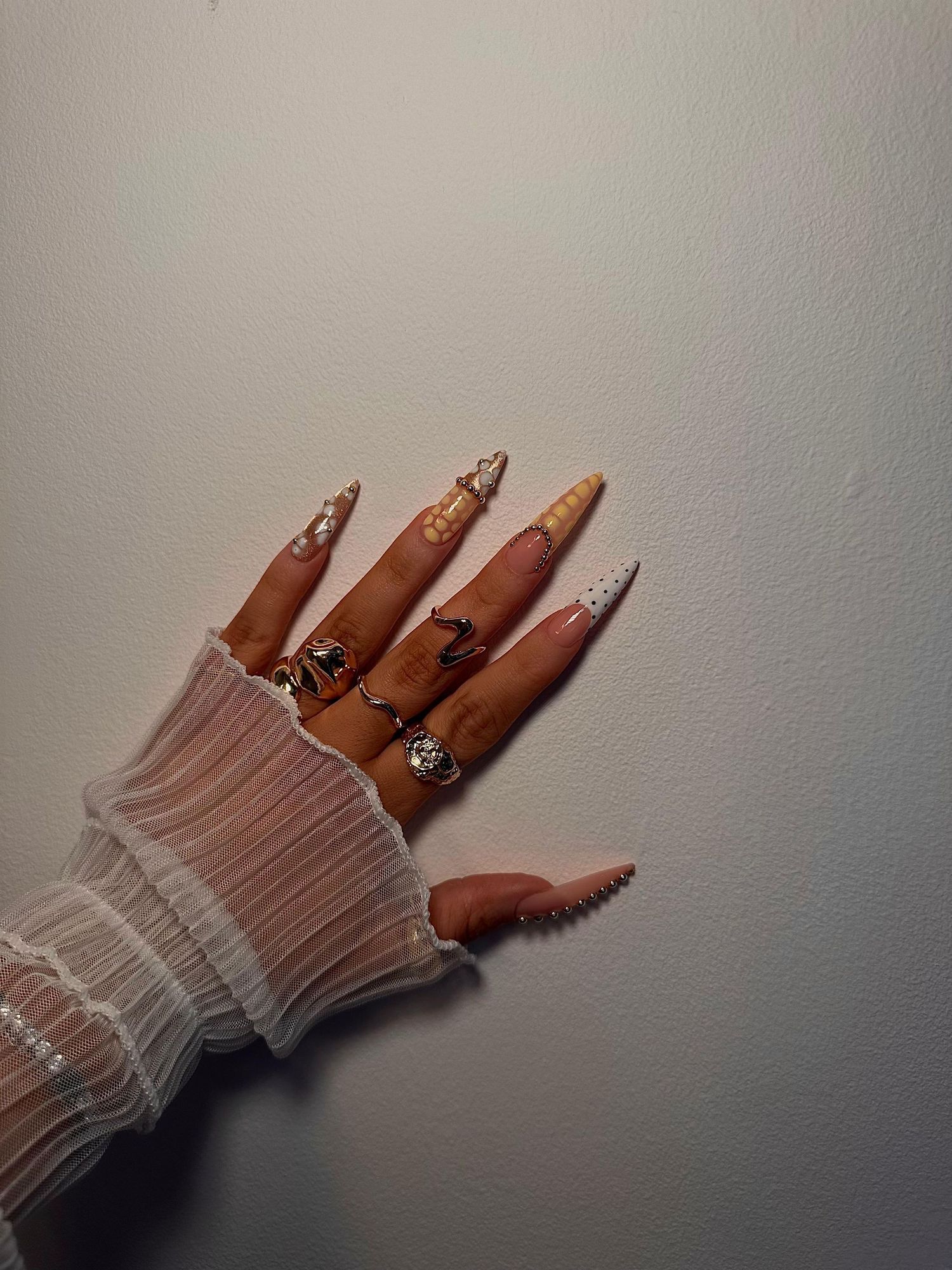Hand with long stiletto nails featuring pearl and crystal nail art, adorned with silver rings, wearing a white lace sleeve.