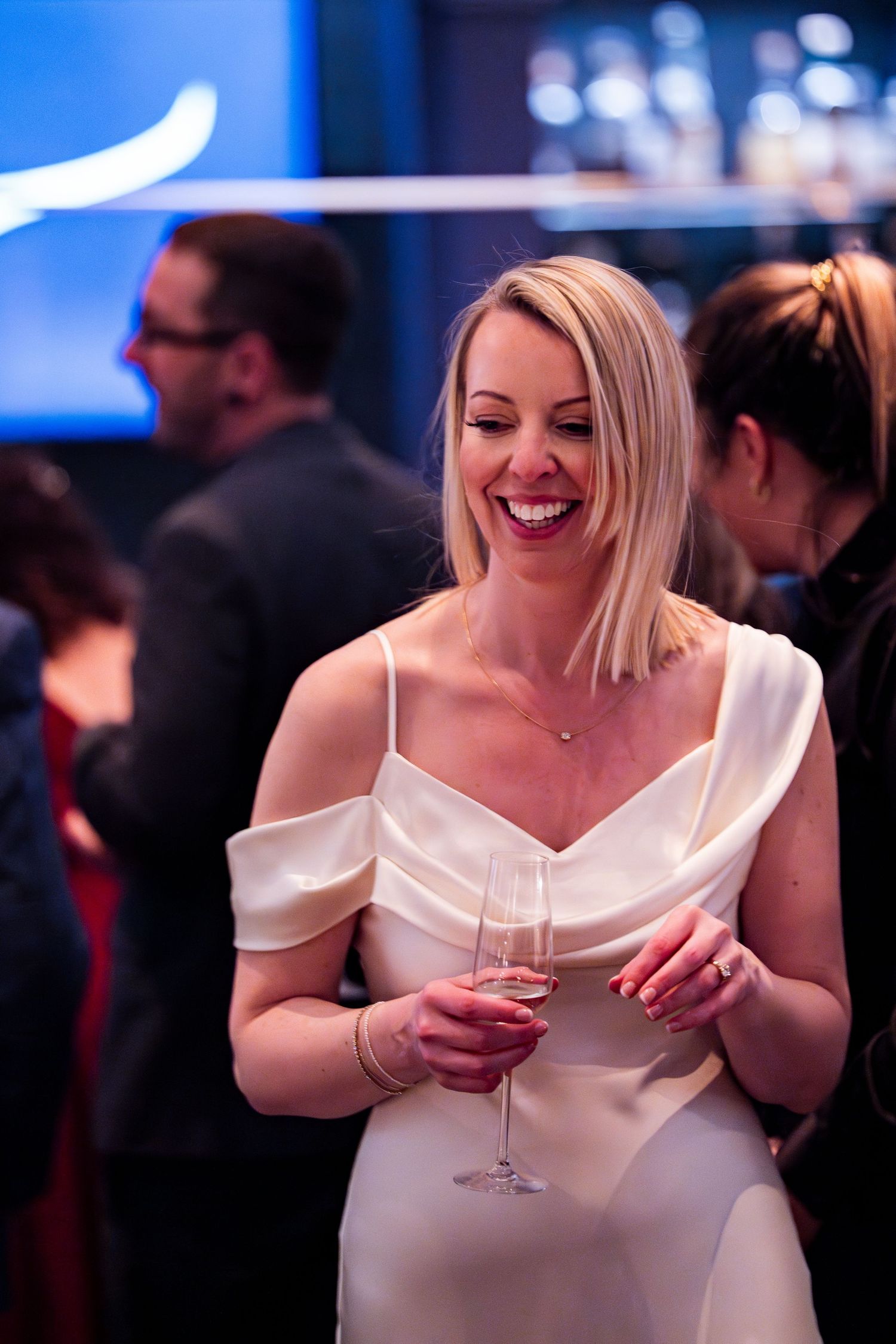 Smiling blonde woman in white off-shoulder dress holding champagne glass at elegant social event.