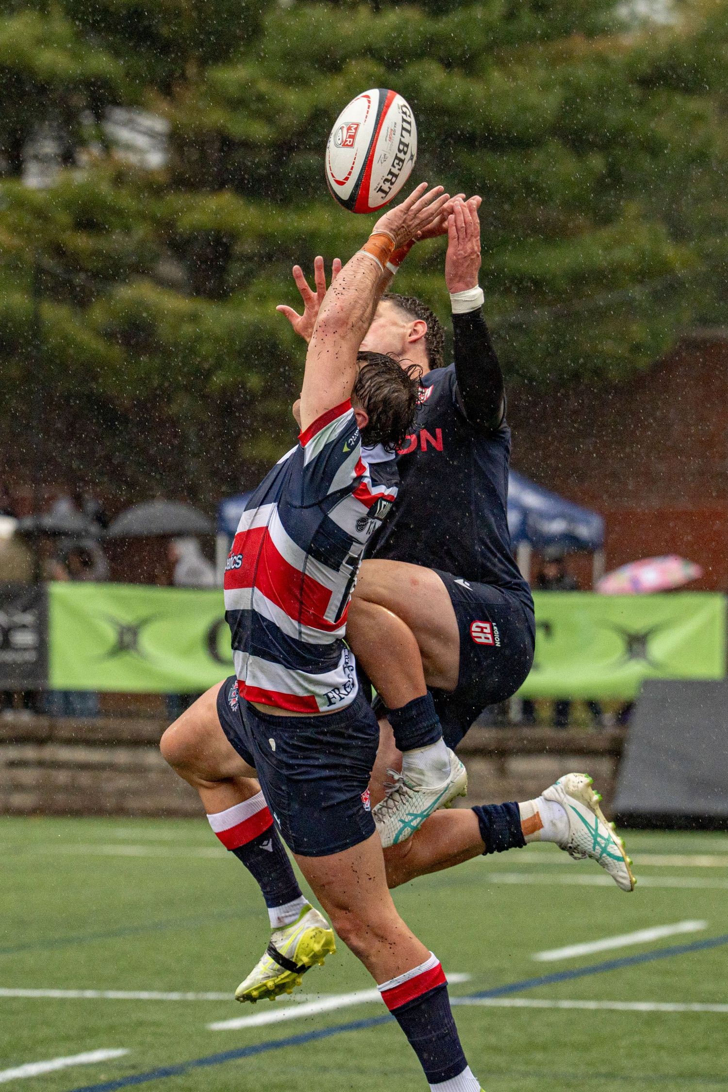 Two rugby players jump to contest a high ball in the rain during an intense match.