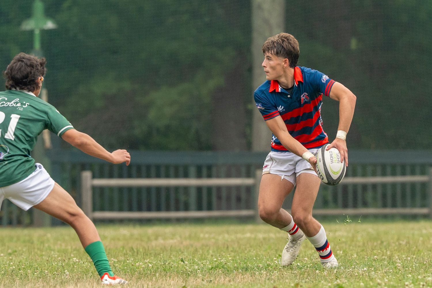 Two rugby players compete on a grass field, one in blue and red holding the ball, the other in green defending.