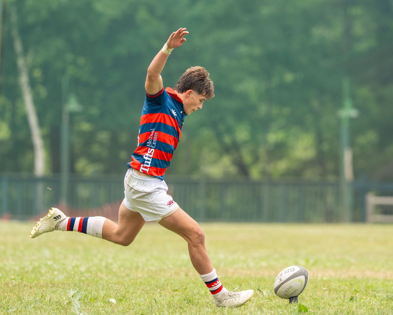 Young rugby player in red and blue striped jersey kicking ball on green grass field during match.
