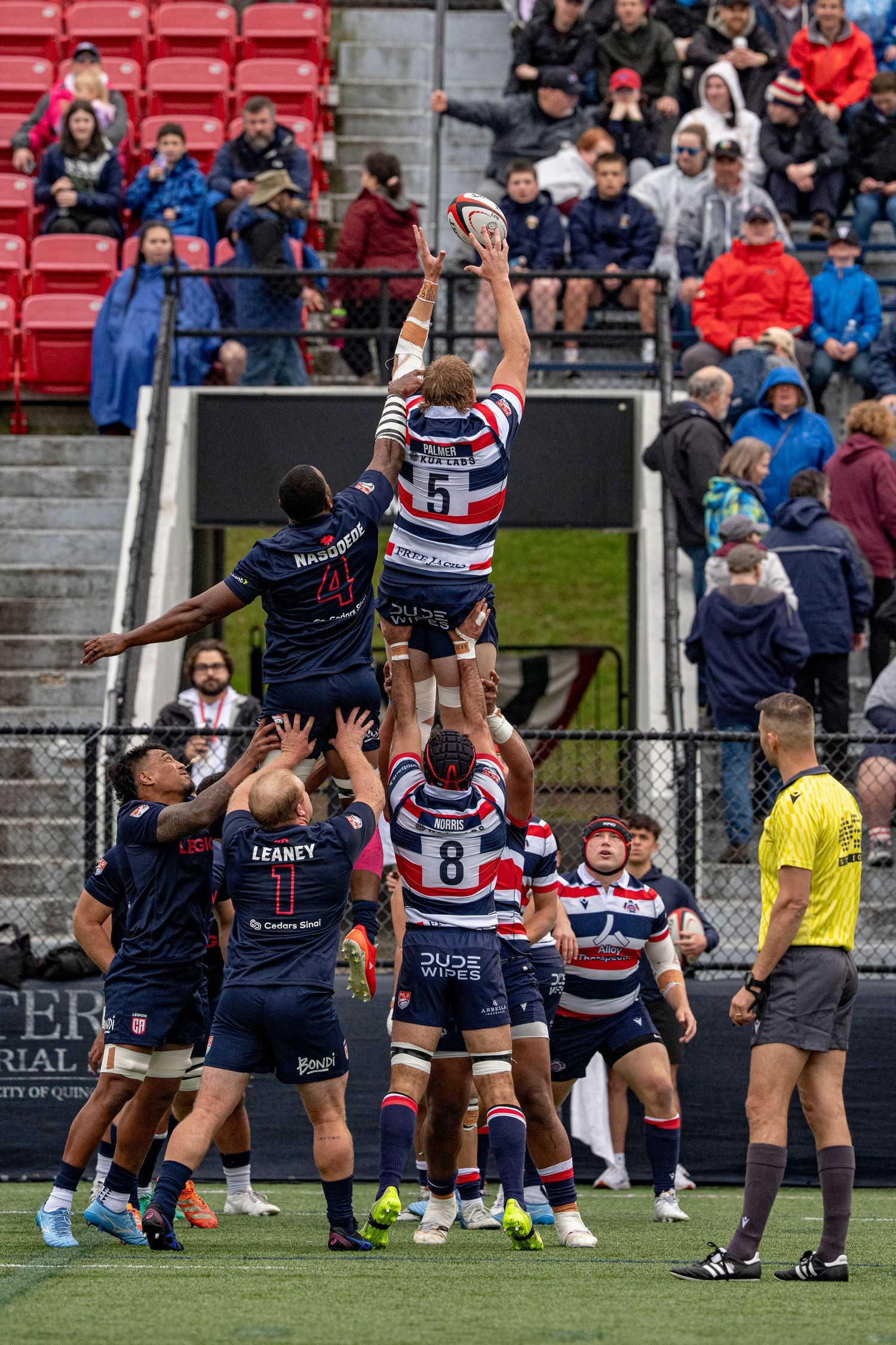 Rugby lineout with a player lifted high to catch the ball as spectators watch from the stands.