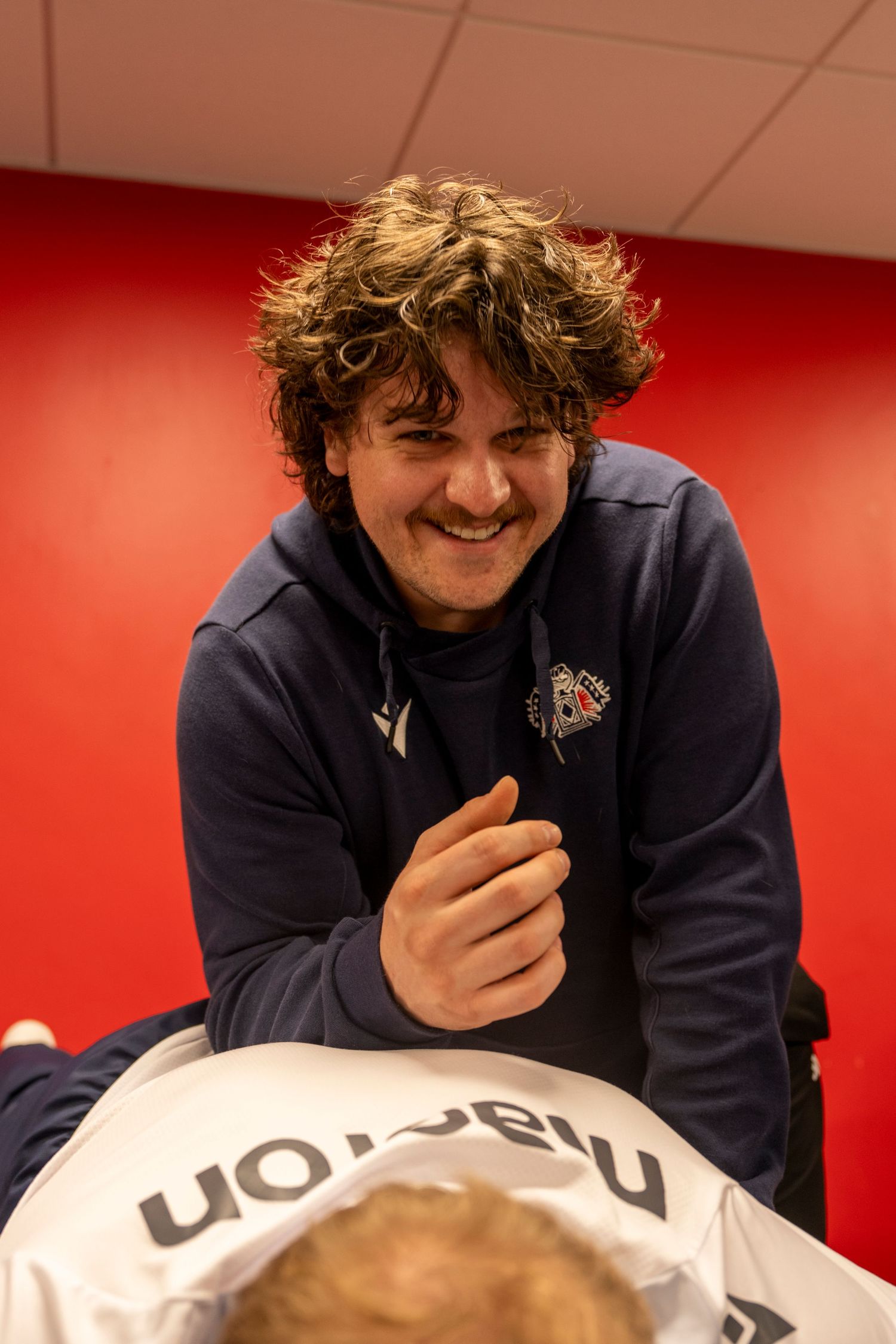 Smiling young man in navy team hoodie leaning over treatment table in locker room.