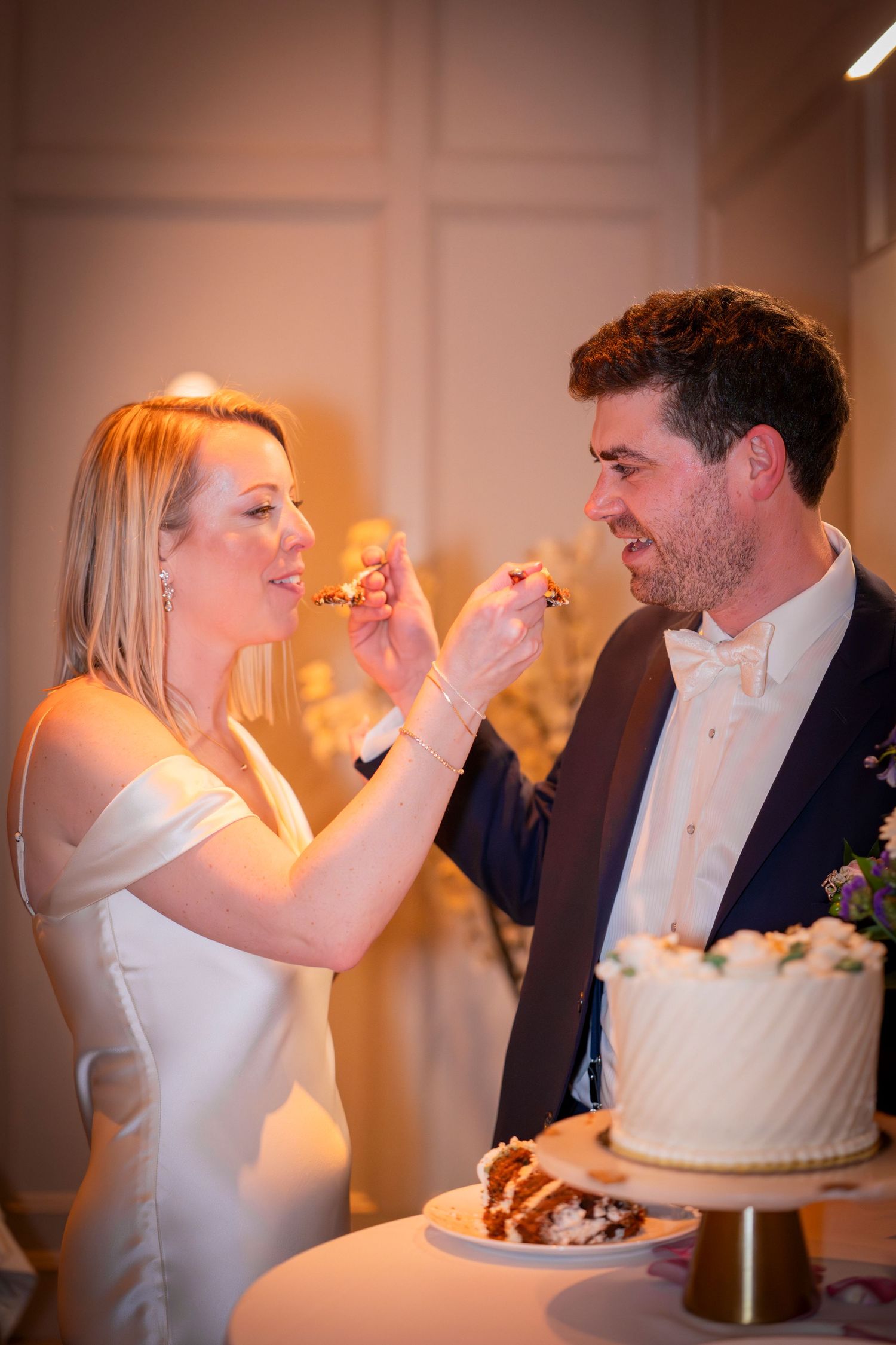 Bride and groom feed each other wedding cake at their elegant indoor reception celebration.