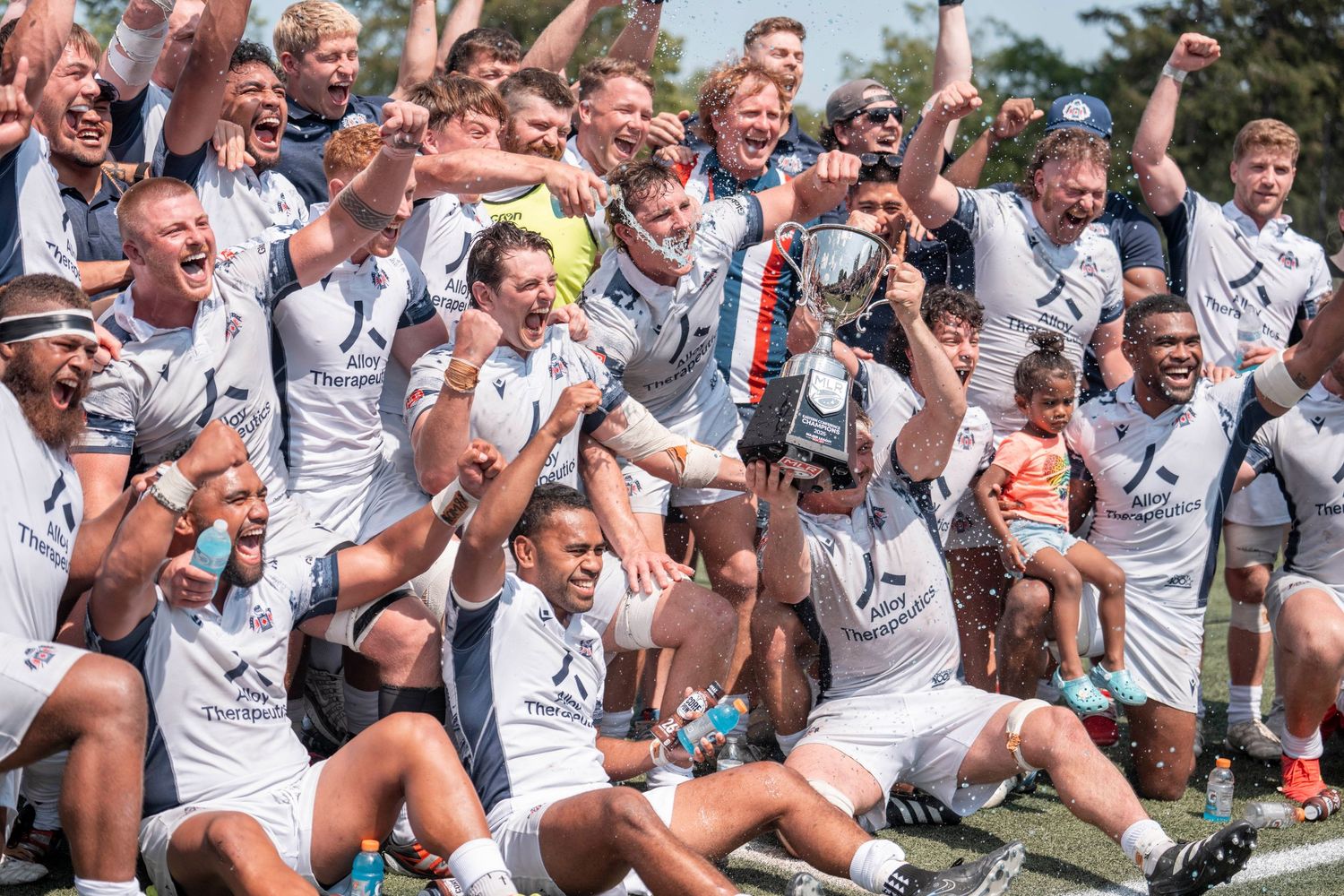 Victorious rugby team celebrating championship win holding trophy outdoors on sunny day.