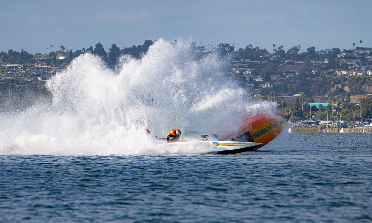 Orange and green speedboat creates a massive water spray while racing across a bay.