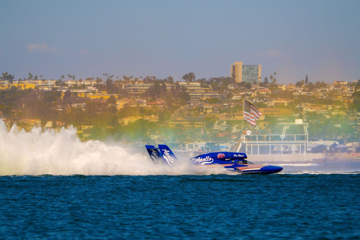 Blue racing powerboat speeds across the water leaving a massive wake near a coastal city.