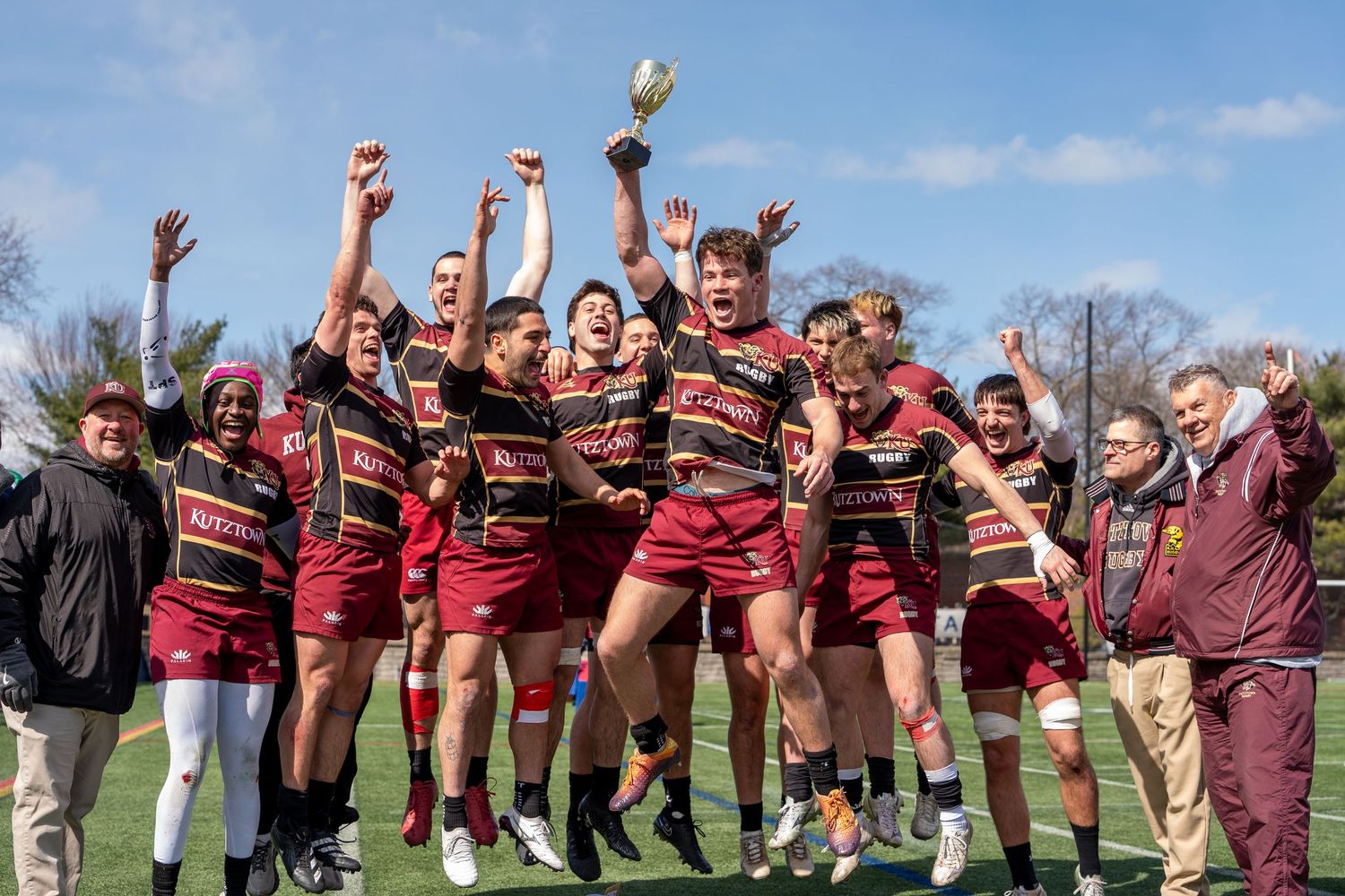 Rugby team celebrates championship victory outdoors on a sunny day, holding trophy aloft.