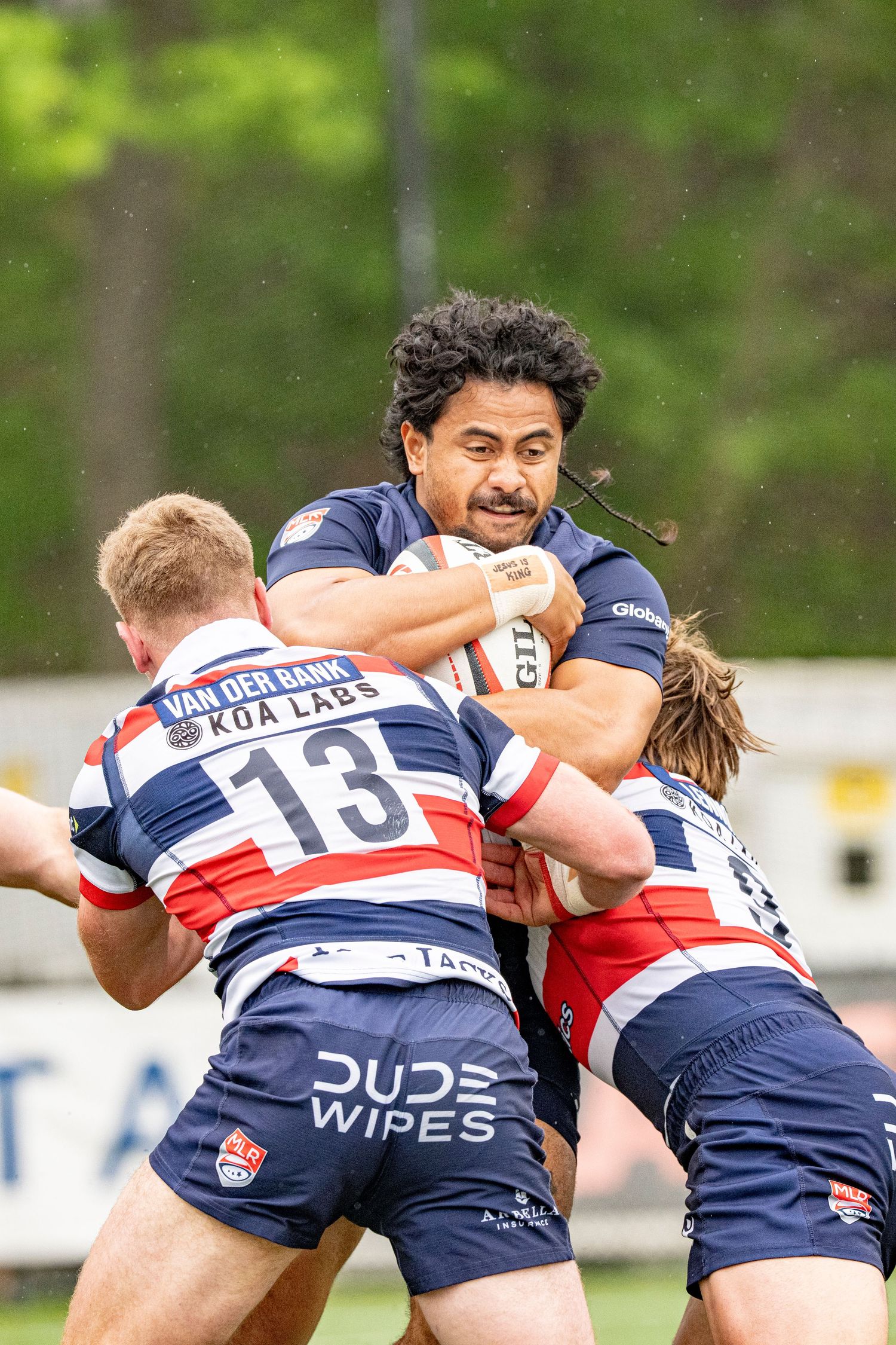 A rugby player powers through two defenders during a rainy match wearing Dude Wipes sponsored jerseys.