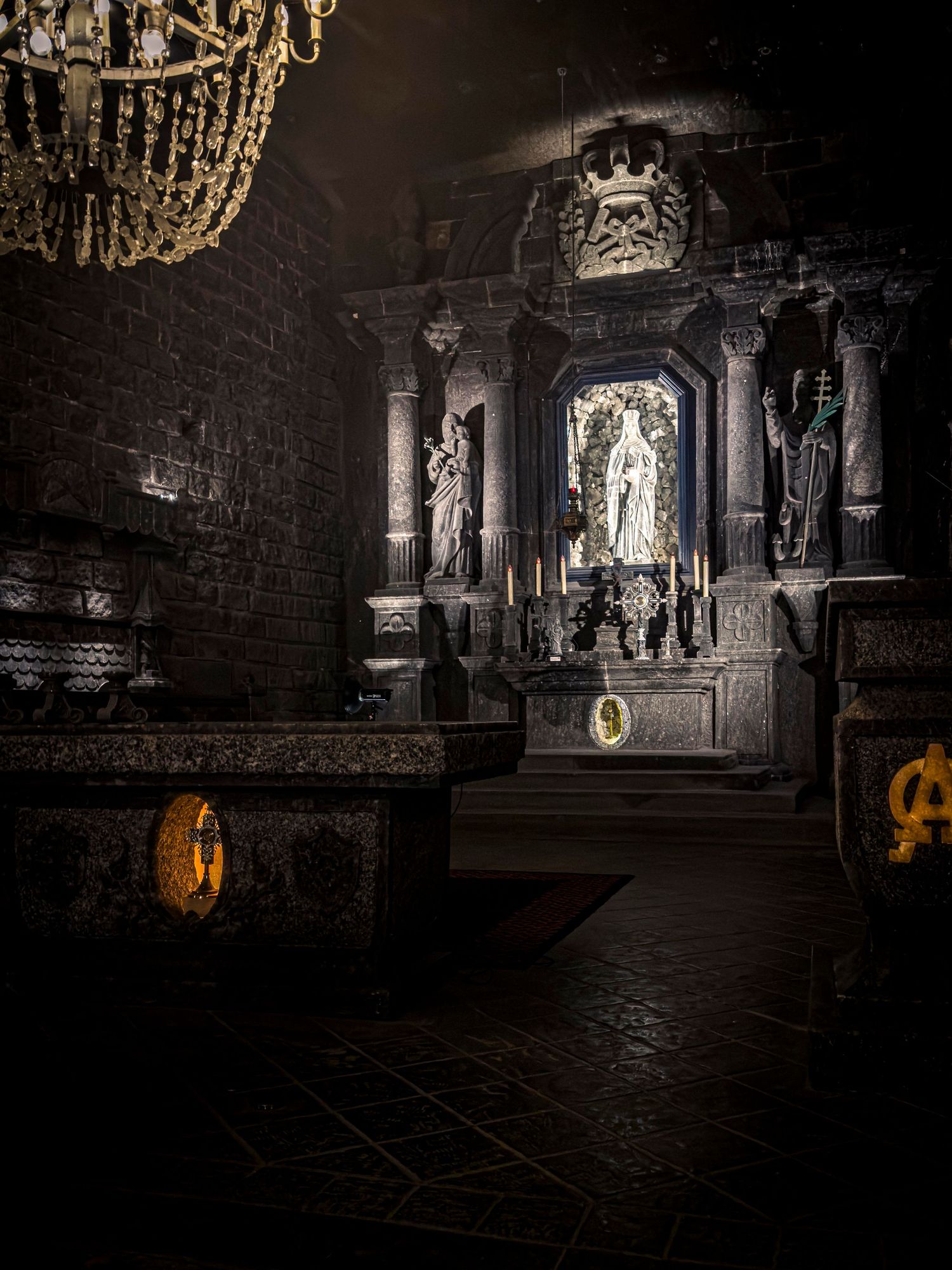 Dark ornate chapel interior with glowing altar, candles, and crystal chandelier in Wieliczka Salt Mine.