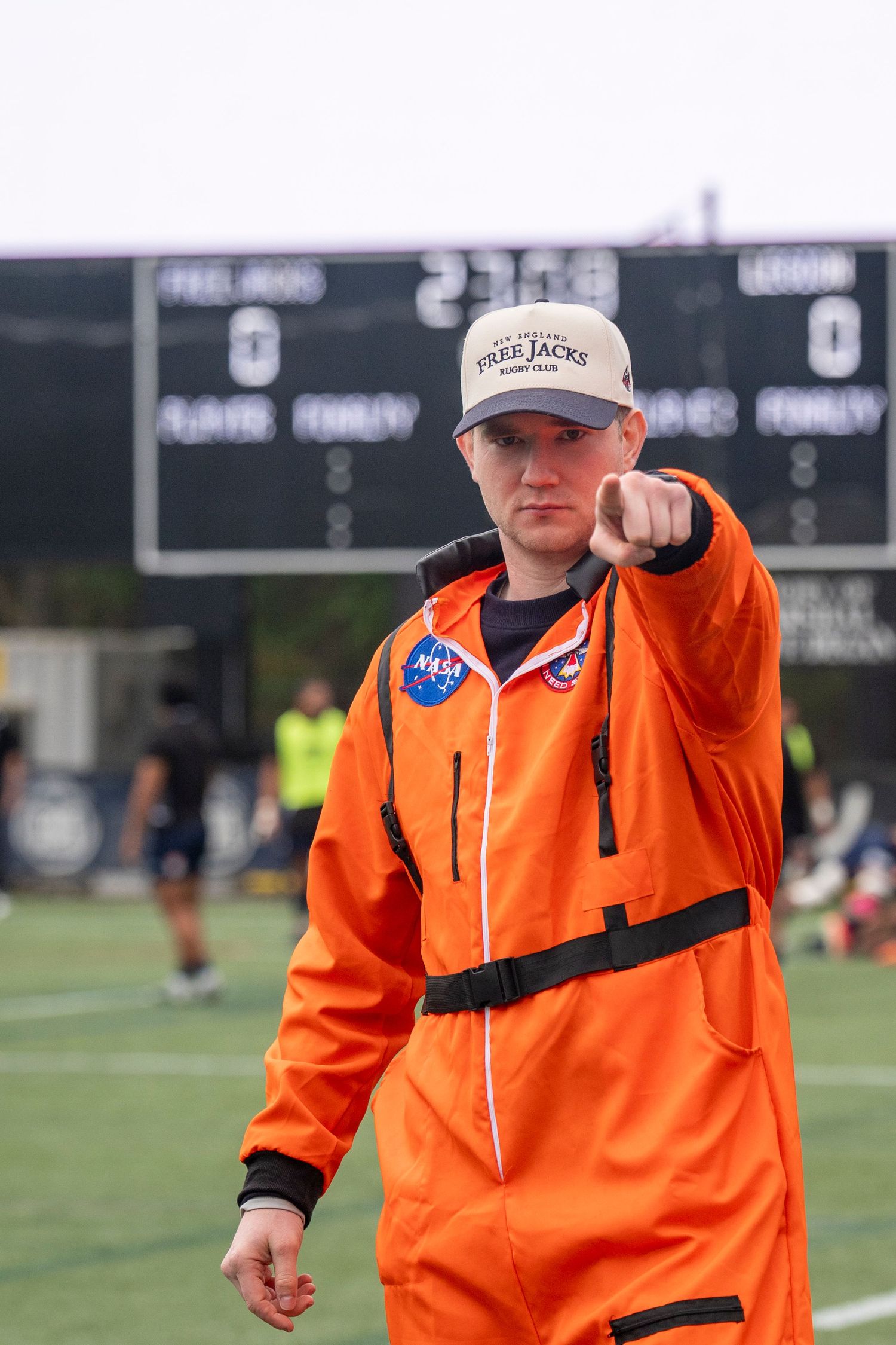 Young fan in NASA orange spacesuit points at camera at a New England Free Jacks rugby game.