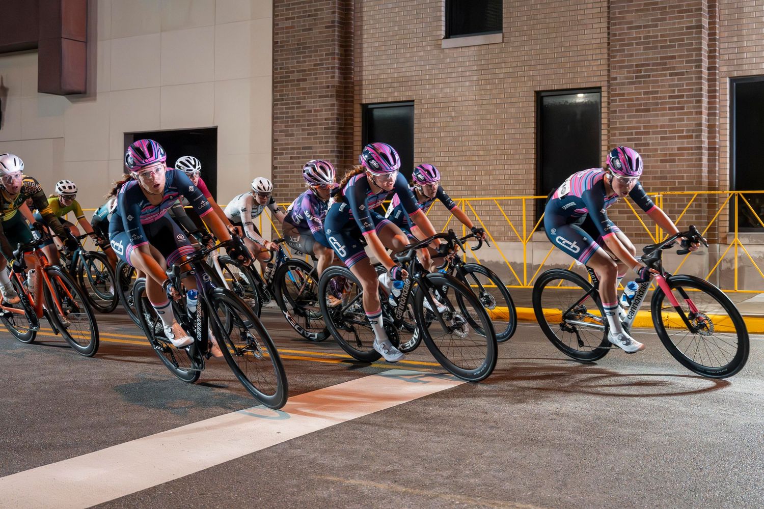 Cyclists in purple jerseys race through a lit urban street at night during a competitive cycling event.