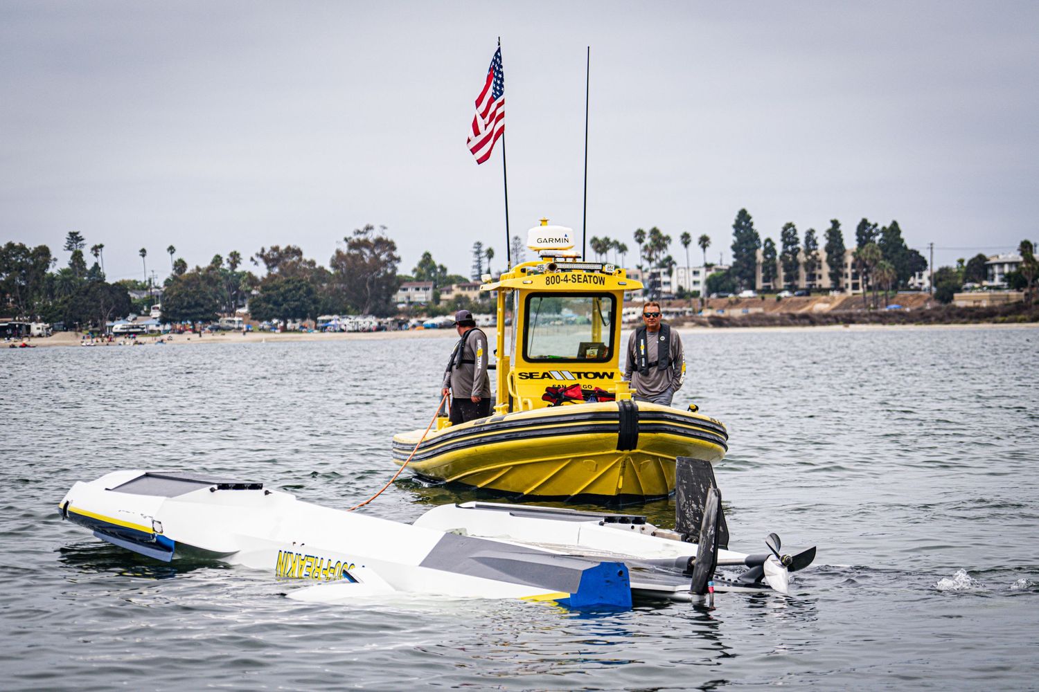 Sea Tow rescue crew responds to capsized white powerboat in calm bay waters under overcast skies.