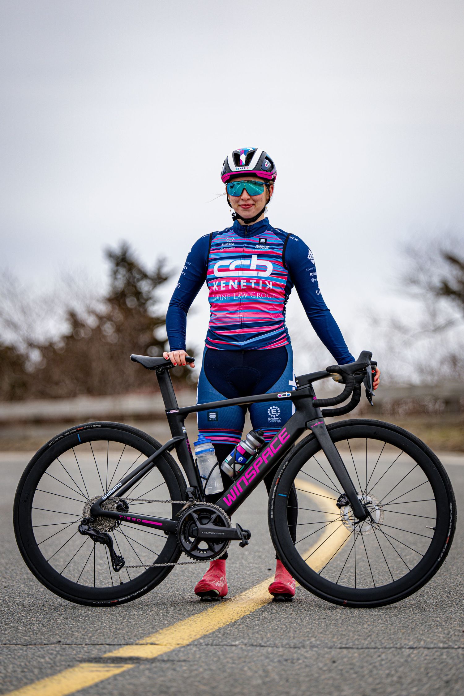 Female cyclist in CCB Kenetix team kit posing with black and pink road bike on pavement.