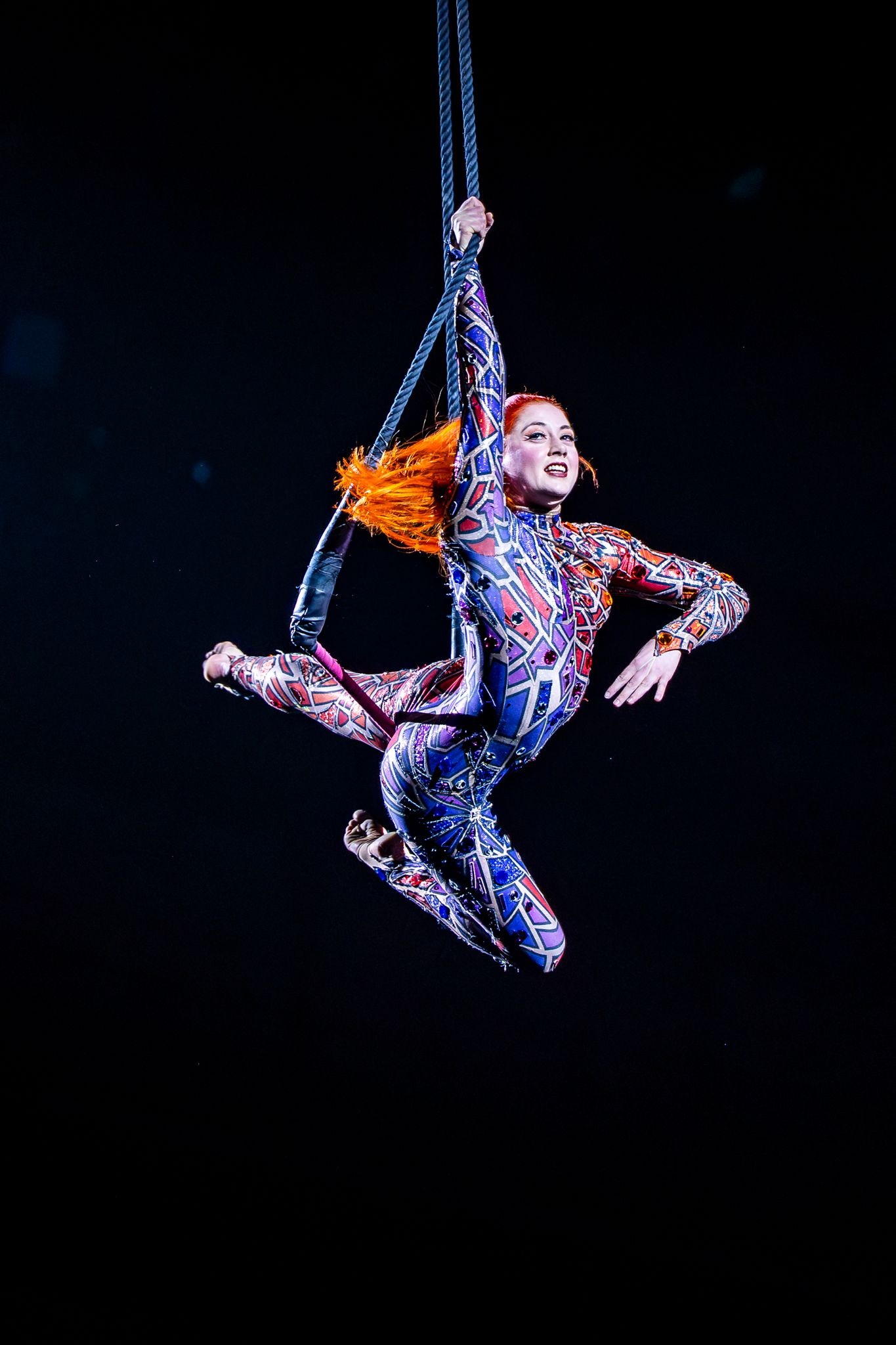 Aerial circus performer in colorful costume suspended on ropes against a dark stage background.