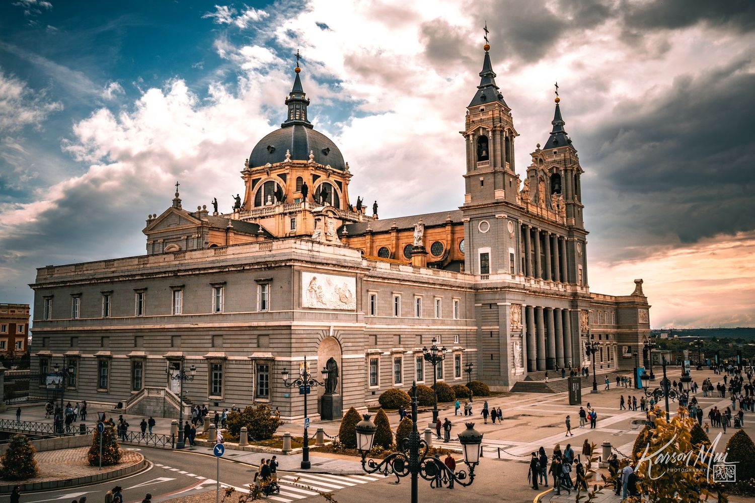 Almudena Cathedral in Madrid stands majestically under dramatic cloudy sky with tourists in plaza.