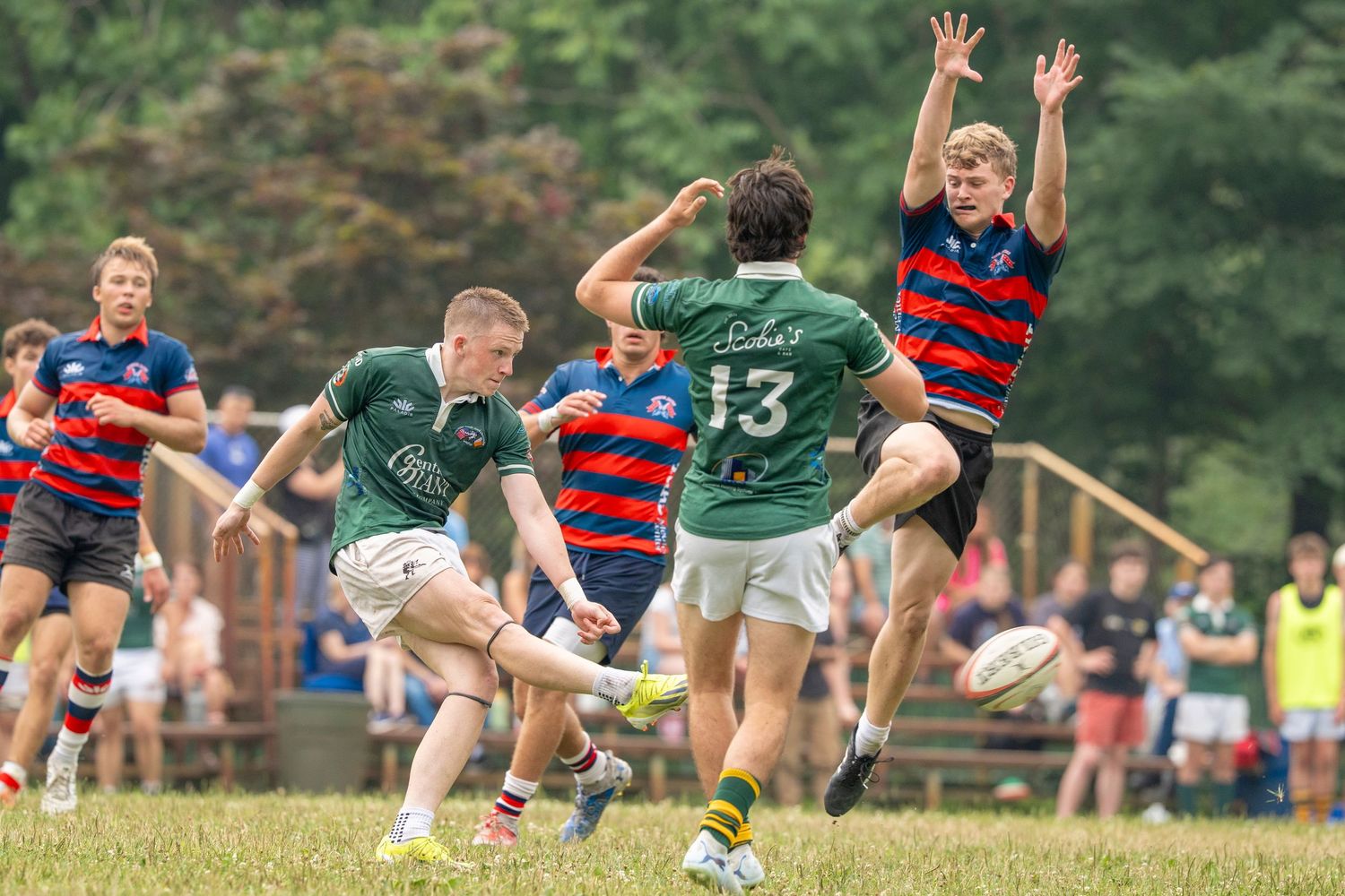Rugby player kicks the ball mid-match while teammates and opponents compete on a grass field.