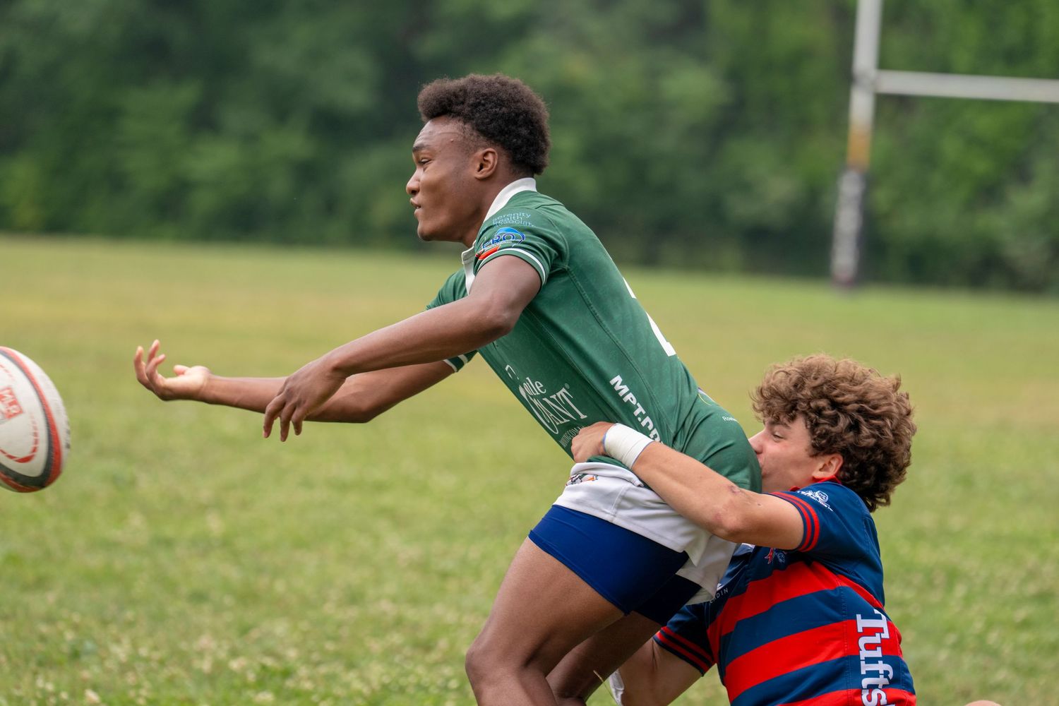 Two youth rugby players compete for the ball during an outdoor match, one in green and one in red and blue.