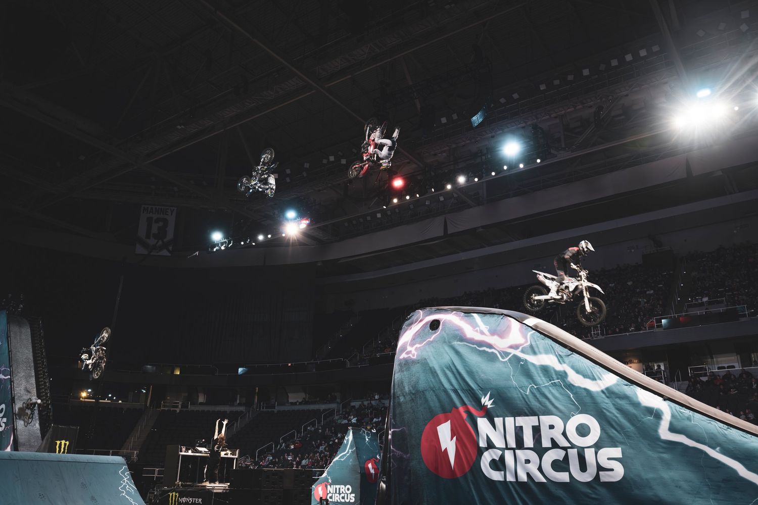 Motocross riders perform aerial tricks over a Nitro Circus ramp at an indoor arena.