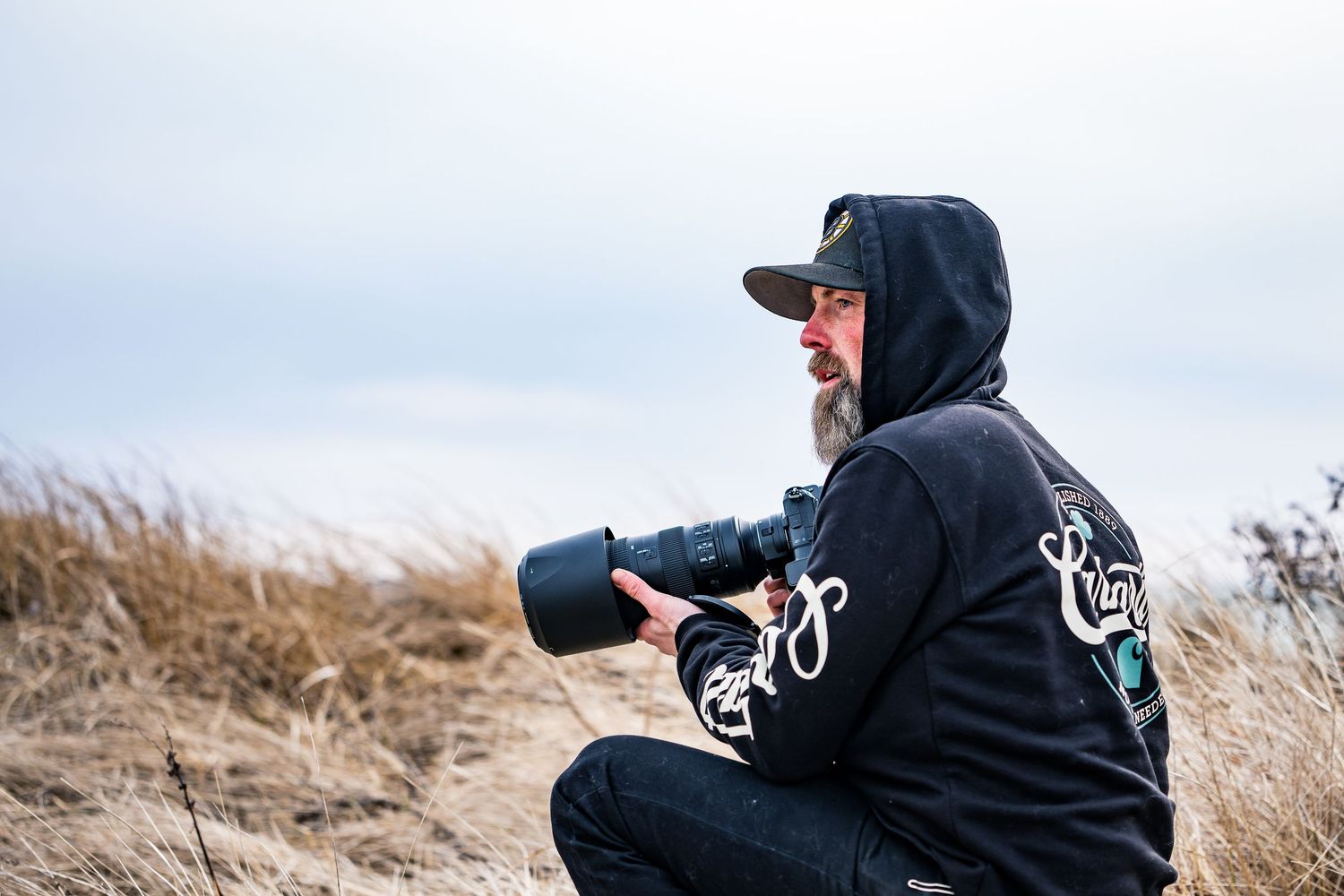 Bearded photographer crouching in tall grass holding professional camera with telephoto lens.