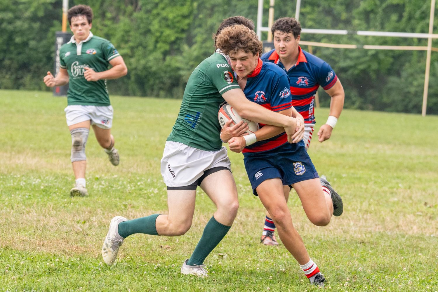 Rugby players compete in a match, with a player in green tackled by opponents in red and blue uniforms on a grass field.