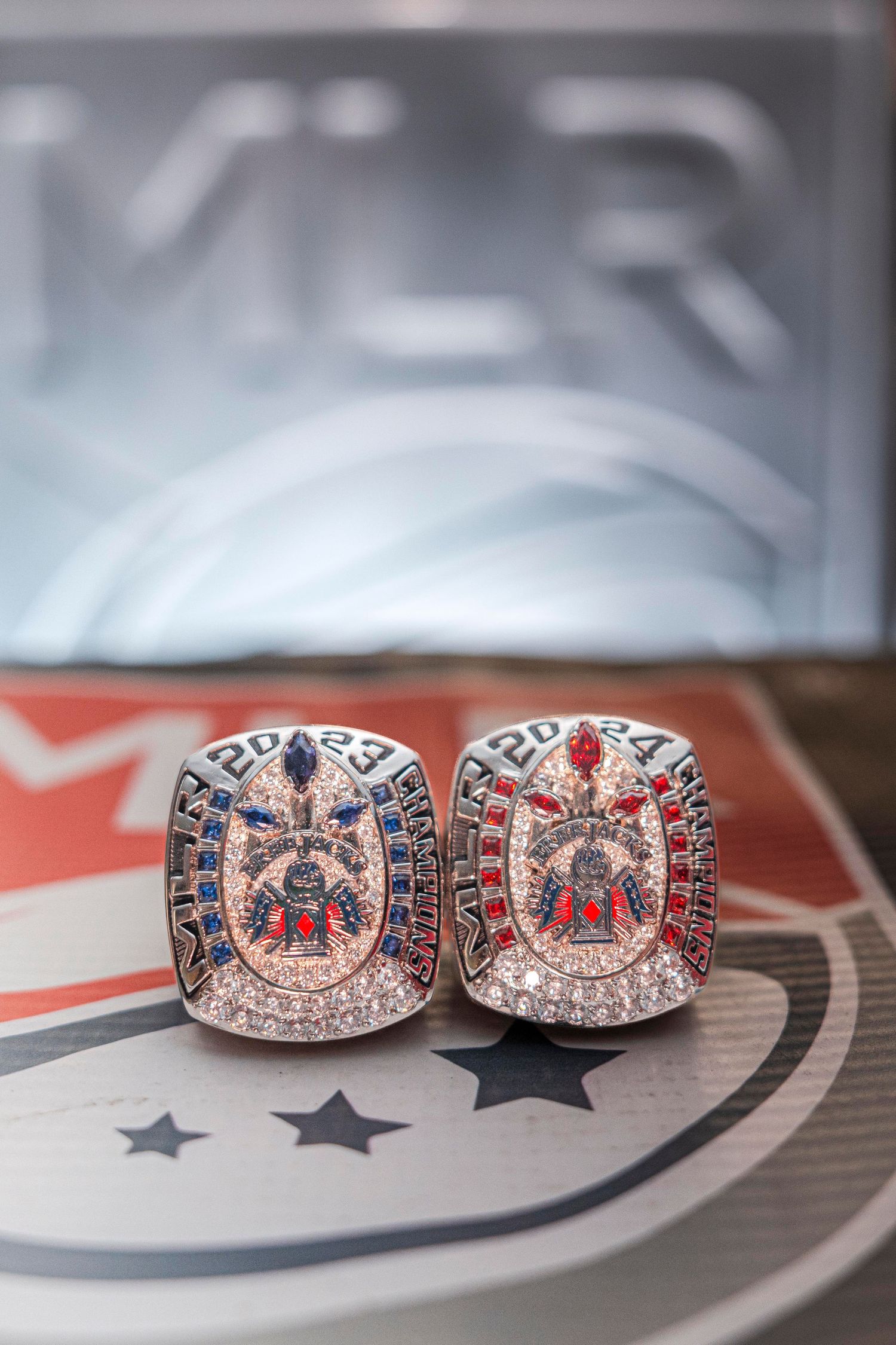 Two MLR championship rings with red and blue gemstones displayed on league logo surface.