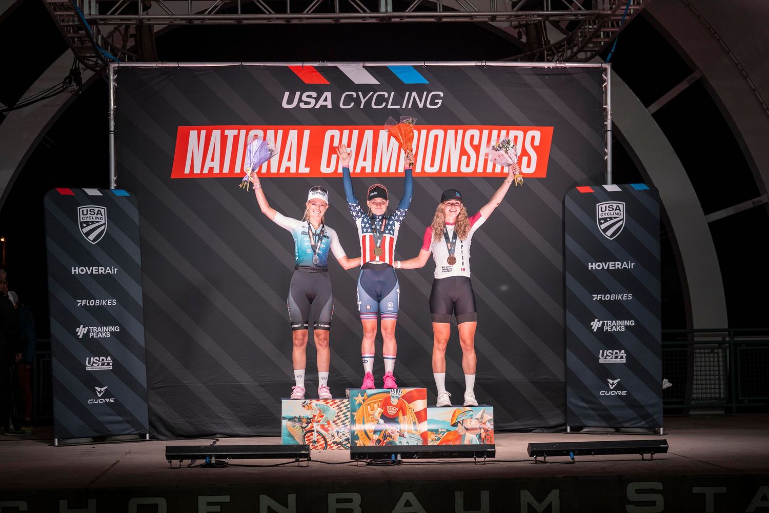 Three cyclists celebrate on the podium at USA Cycling National Championships, with the winner in a stars and stripes jersey.