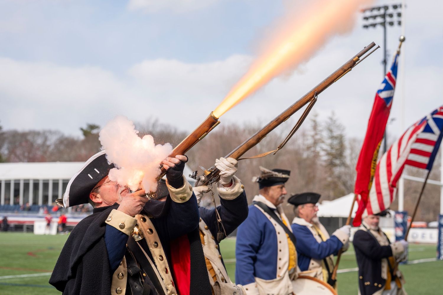 Colonial reenactors fire muskets at a ceremonial event at a sports stadium.