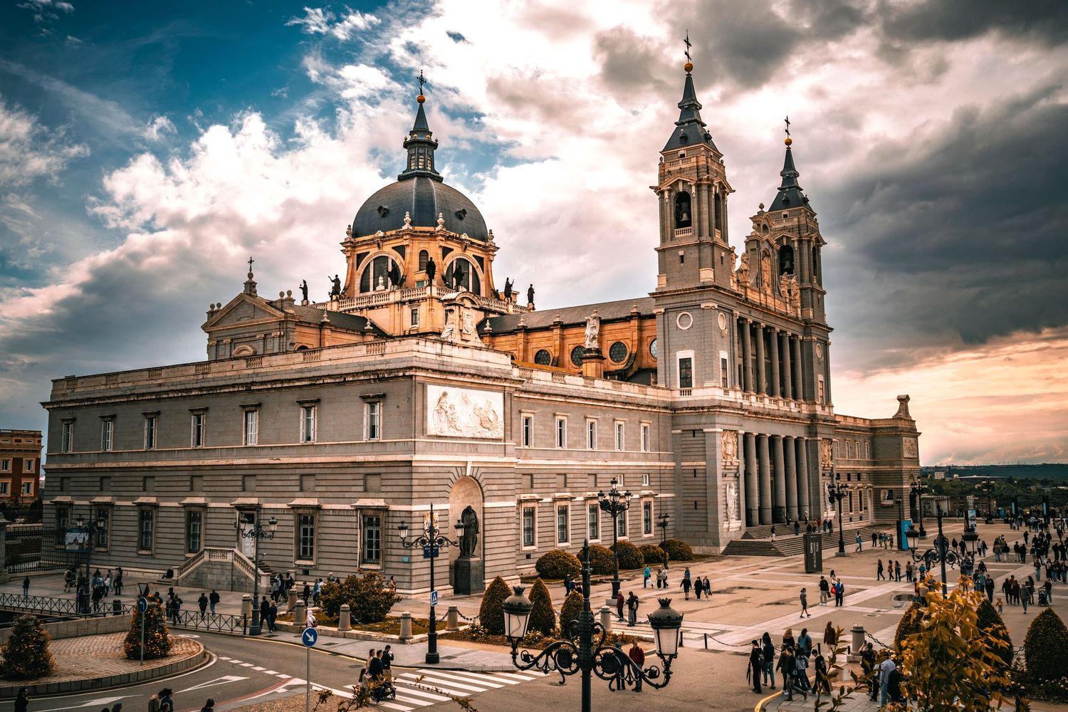 Almudena Cathedral in Madrid, Spain, with dramatic cloudy sky and tourists in the plaza.