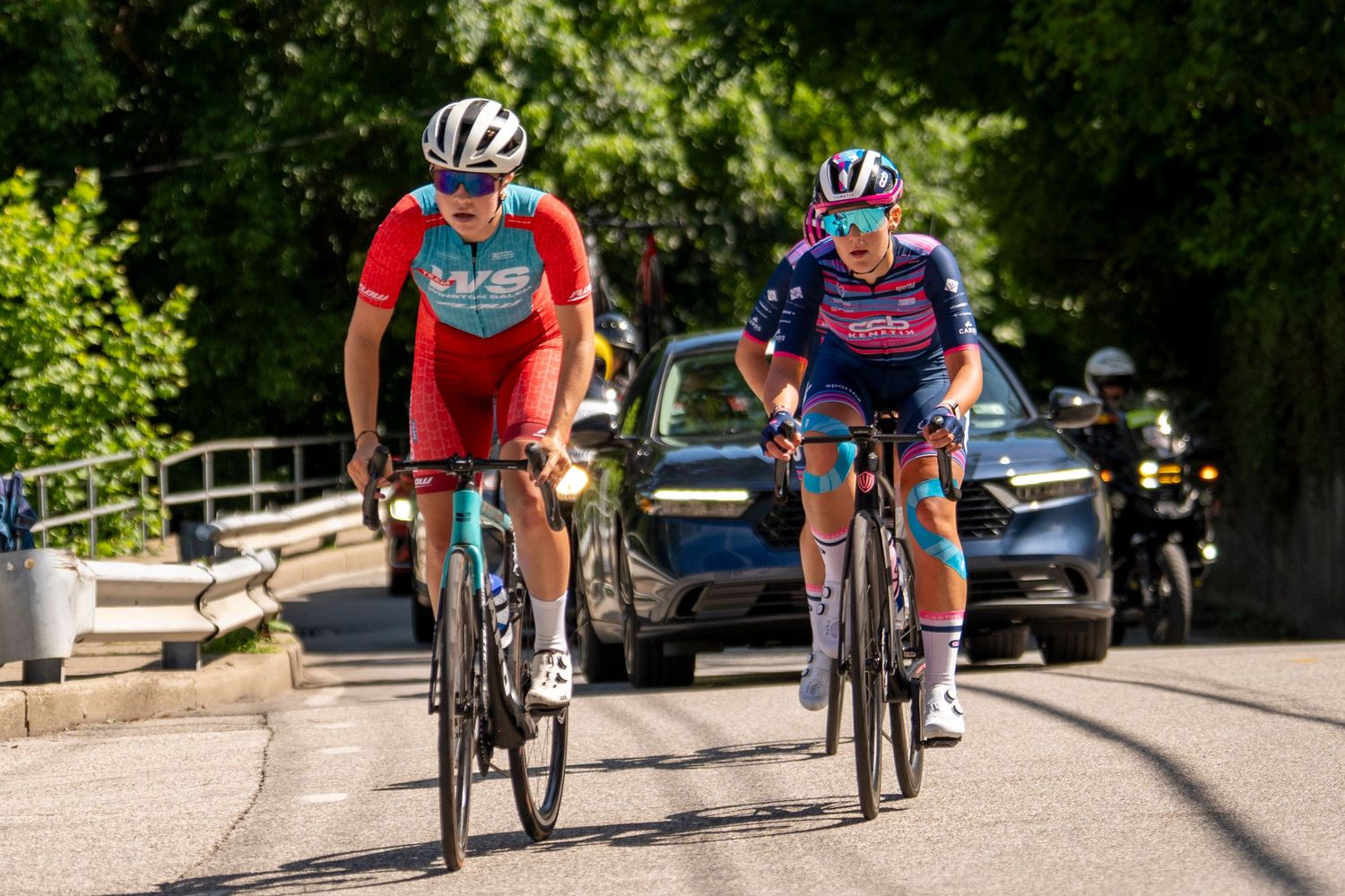 Two cyclists in colorful jerseys and helmets ride road bikes on a tree-lined street during a cycling race.