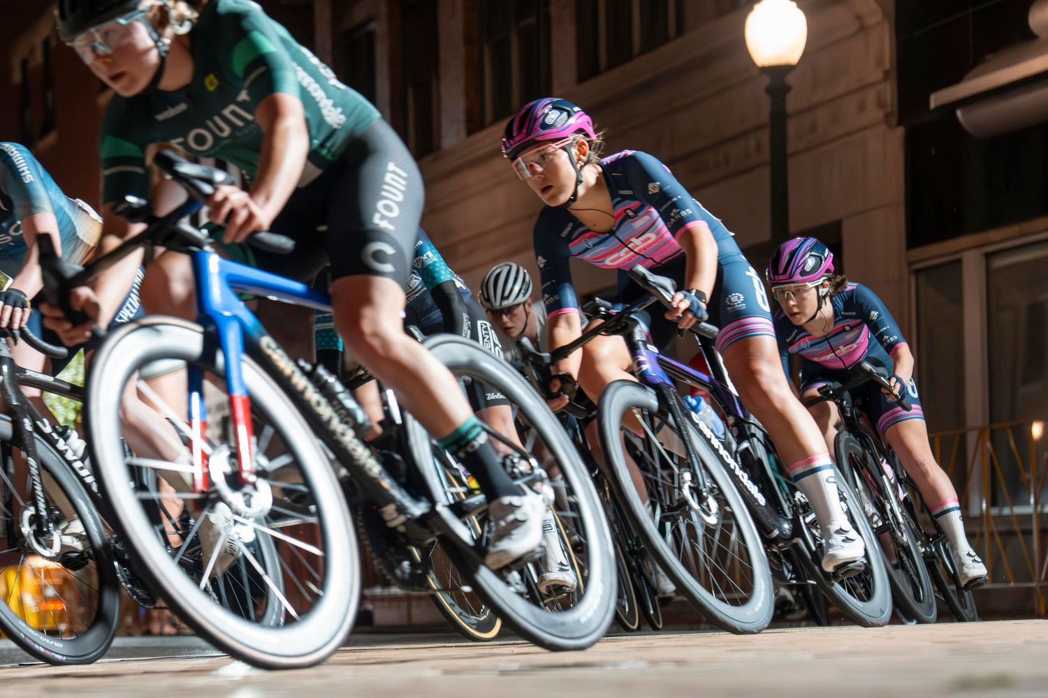 Female cyclists racing at night on a city street, wearing colorful team jerseys, leaning into a turn during a criterium race.