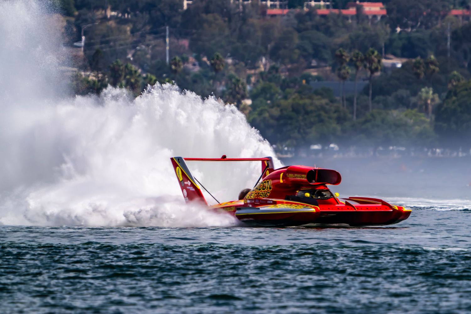 Red and yellow hydroplane racing boat creates massive spray on a sunny day.