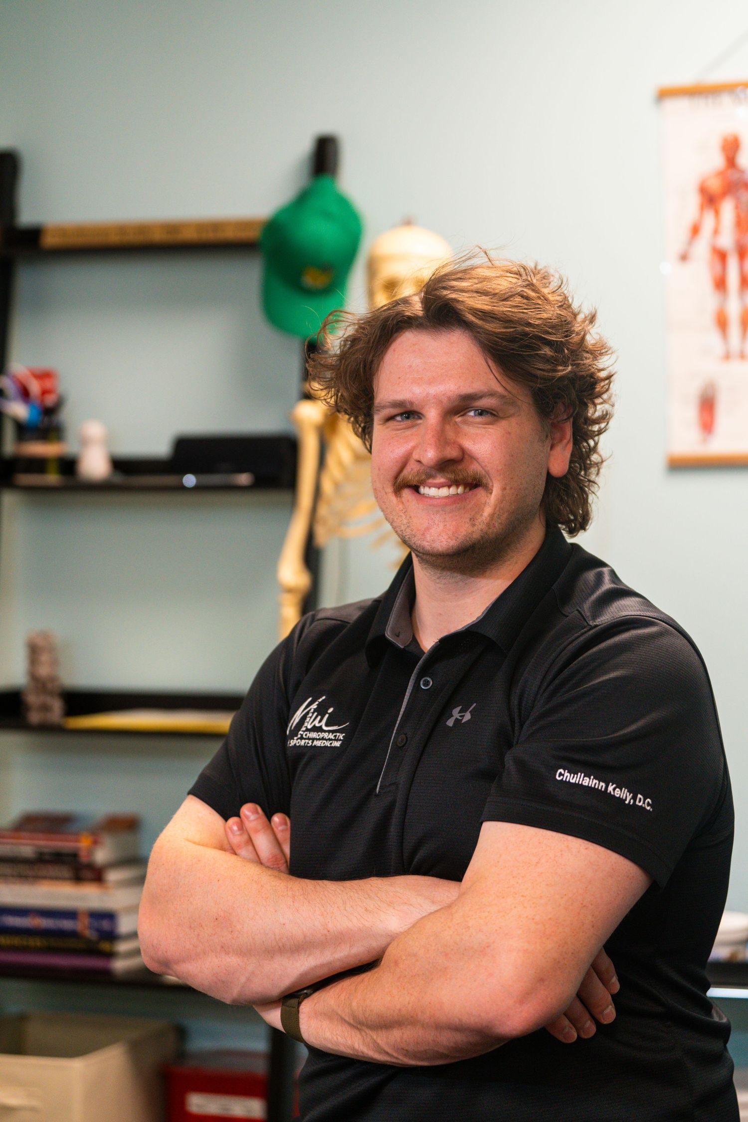 Chiropractor Dr. Kelly smiles with arms crossed in his office, with anatomy poster and skeleton model in background.