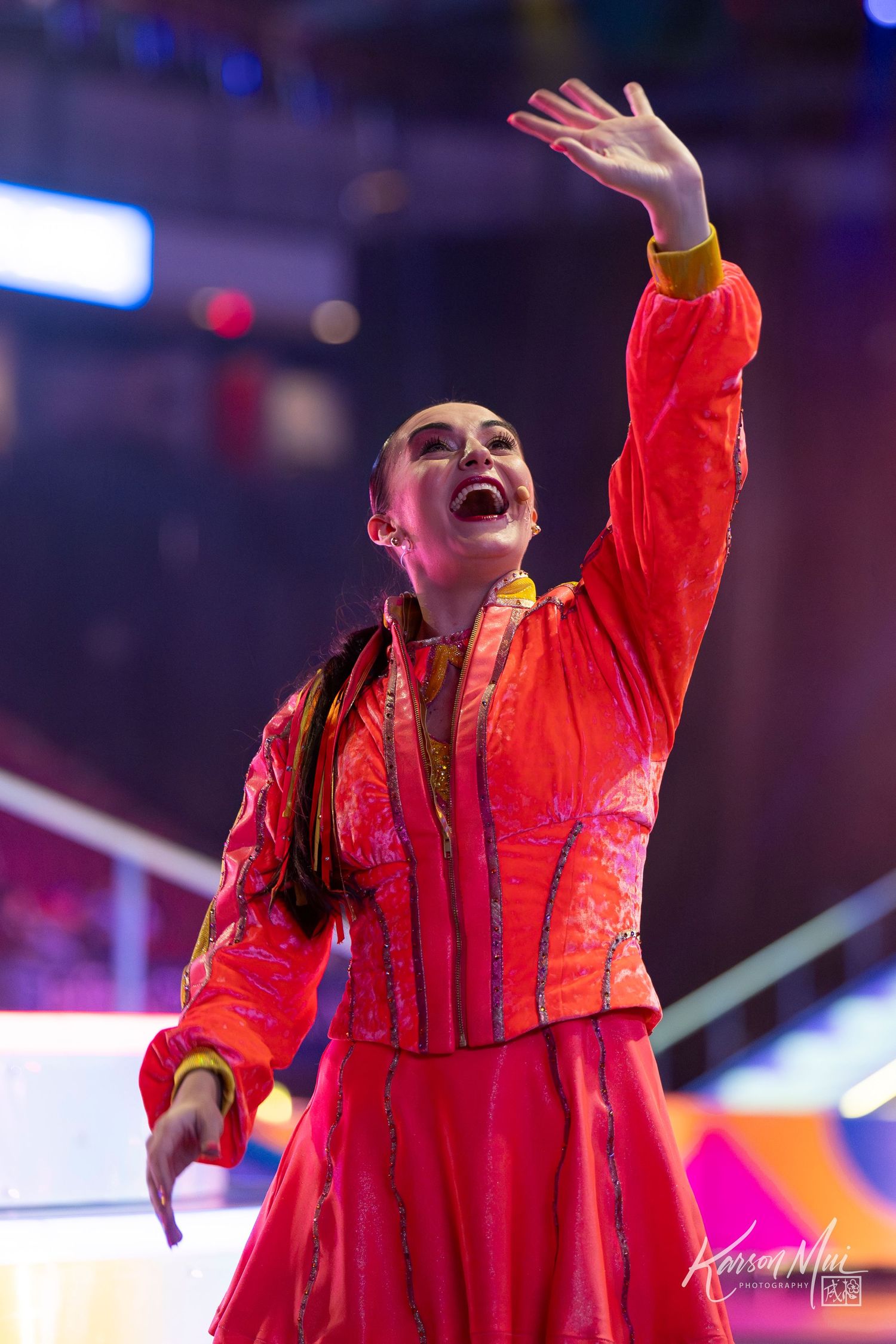 Female performer in vibrant red traditional costume dancing on colorful lit stage.