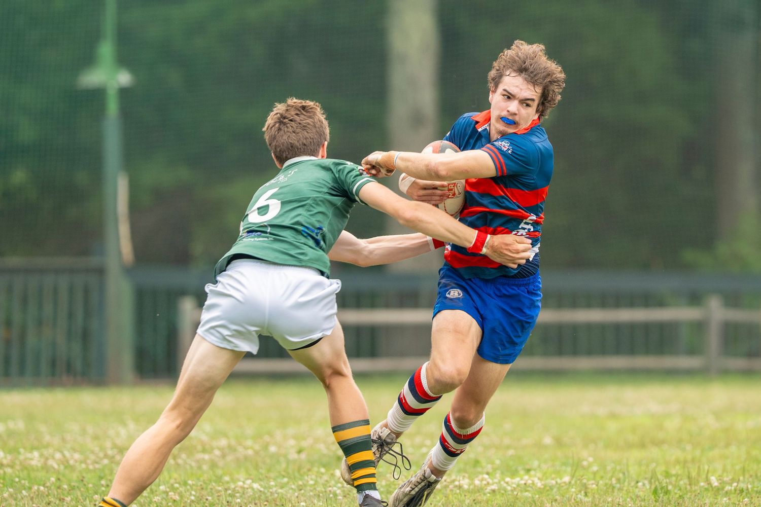 Two young rugby players compete on a grass field, one in green attempting to tackle the other in red and blue stripes.